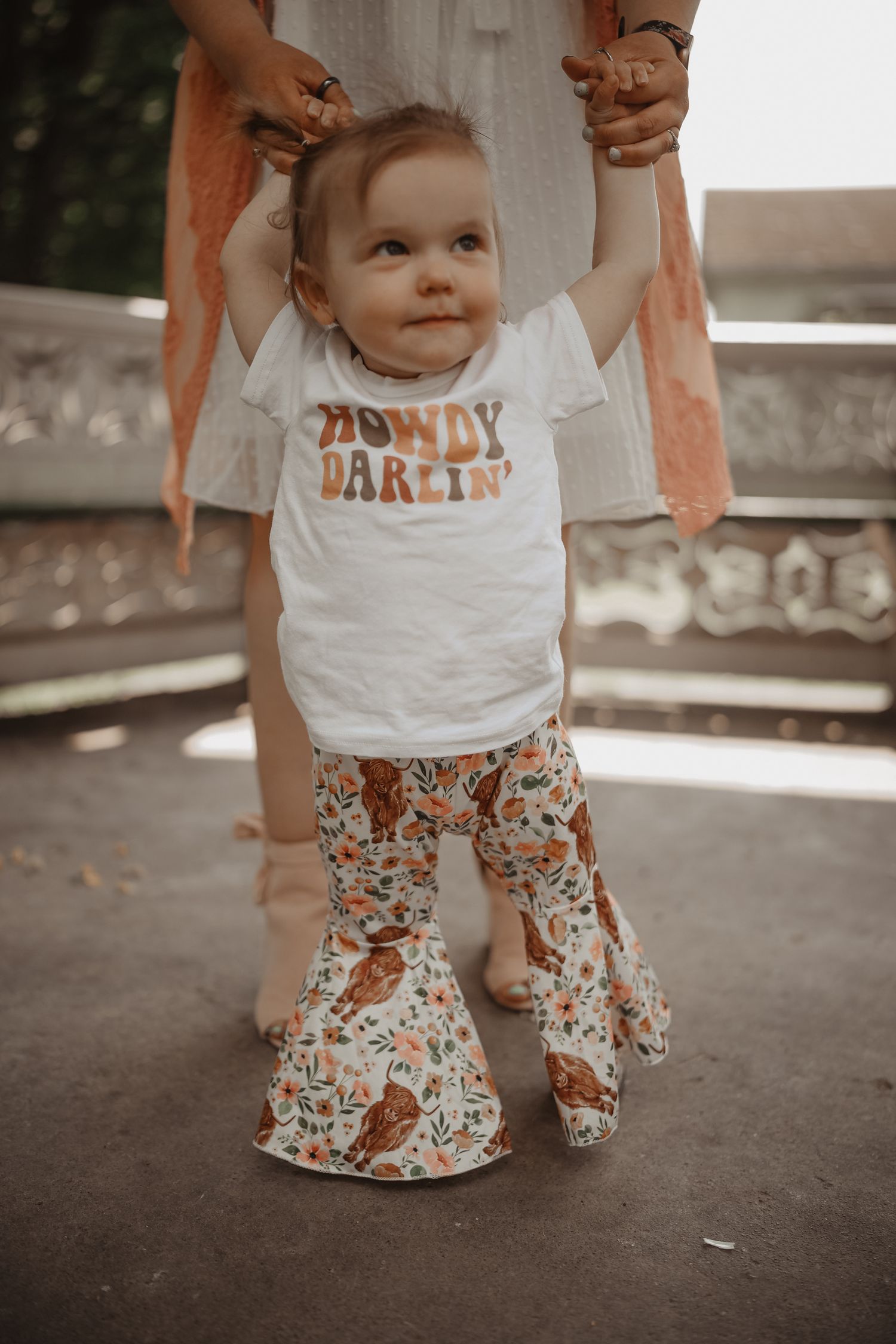 A small child wearing a white t-shirt and floral pants is supported while learning to walk outdoors.