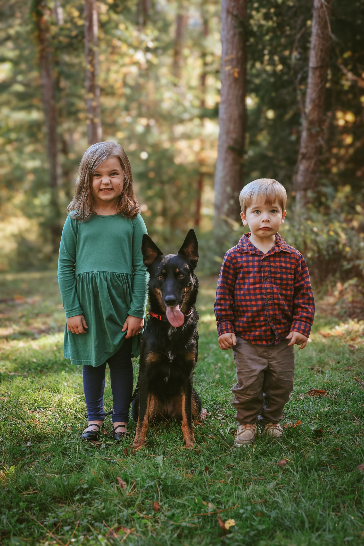 A black and brown doberman stands between two children in casual attire on grass in a wooded setting.