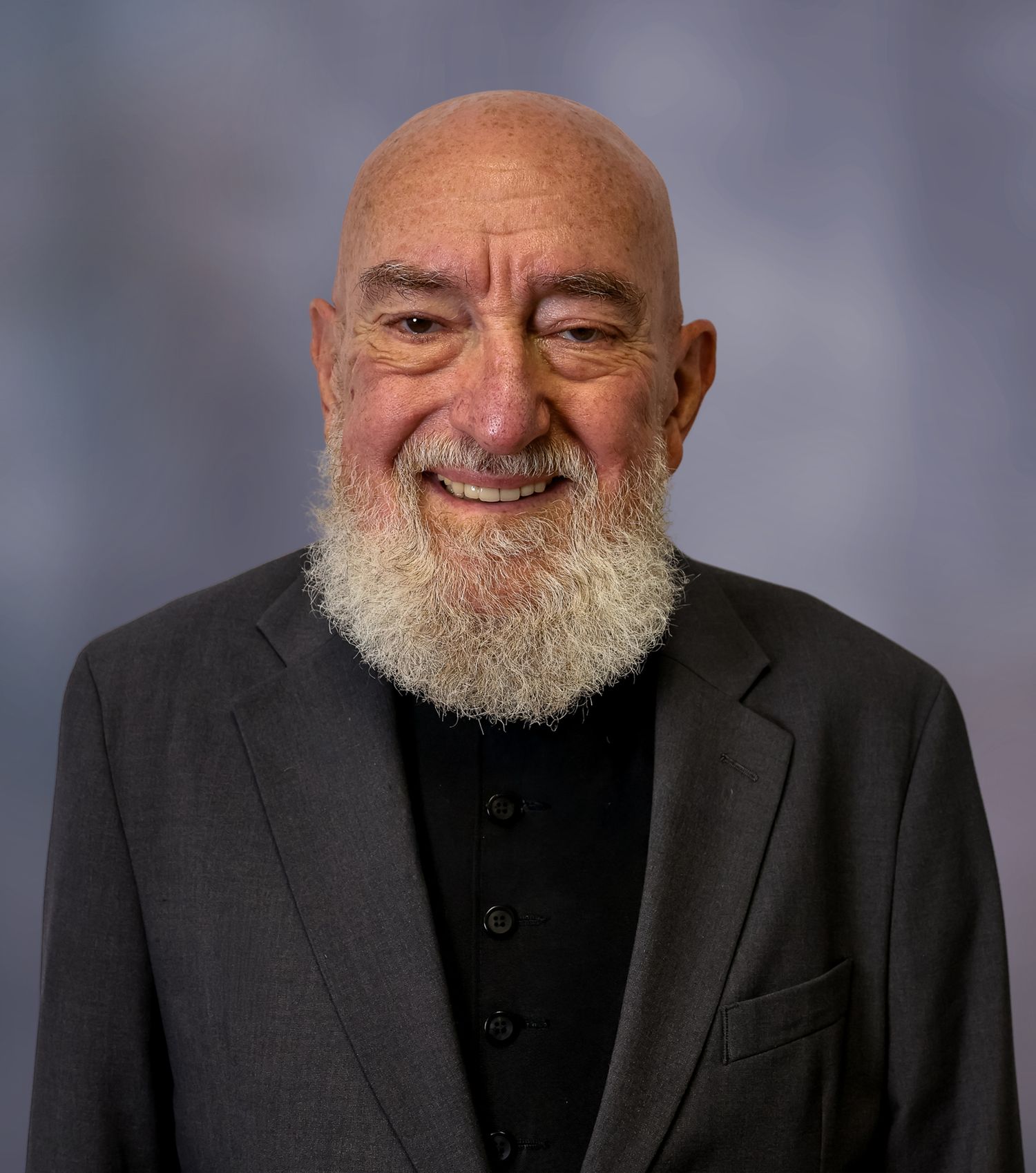 A person with a white beard wearing a black suit jacket smiles warmly in a professional studio portrait series.