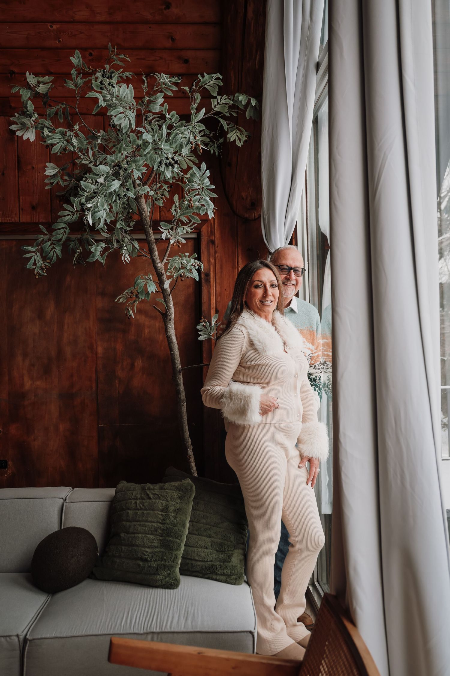 A couple in matching white outfits embraces in front of white curtains and green plants in an indoor setting.