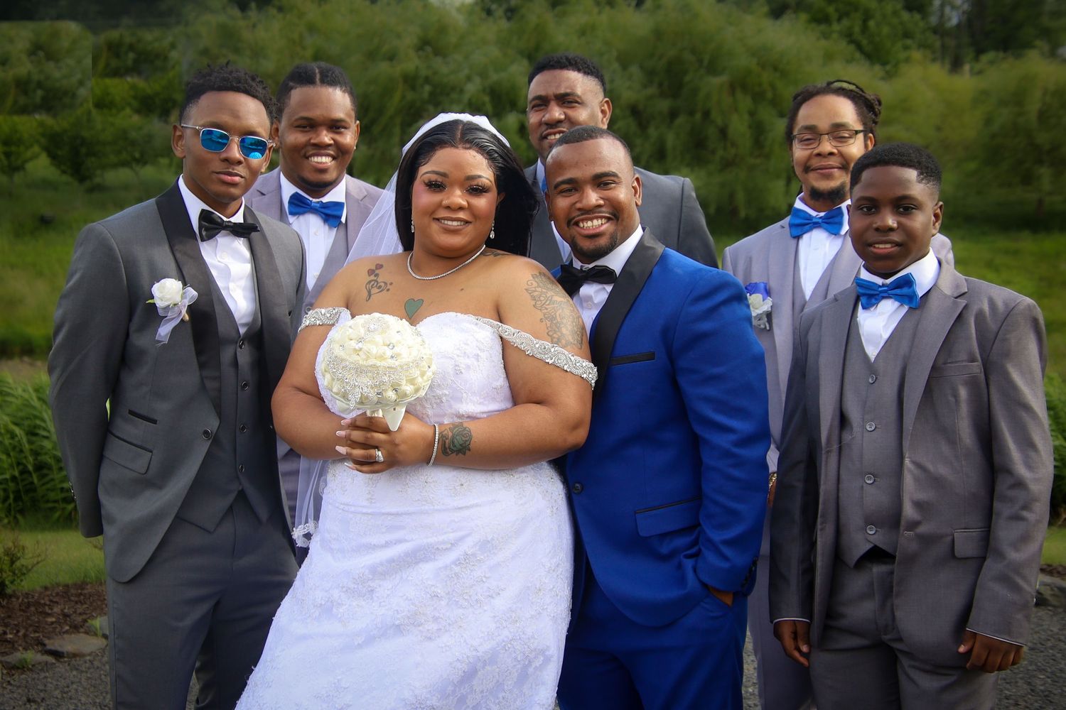 Wedding party members in blue and gray attire pose together outdoors in a garden setting.