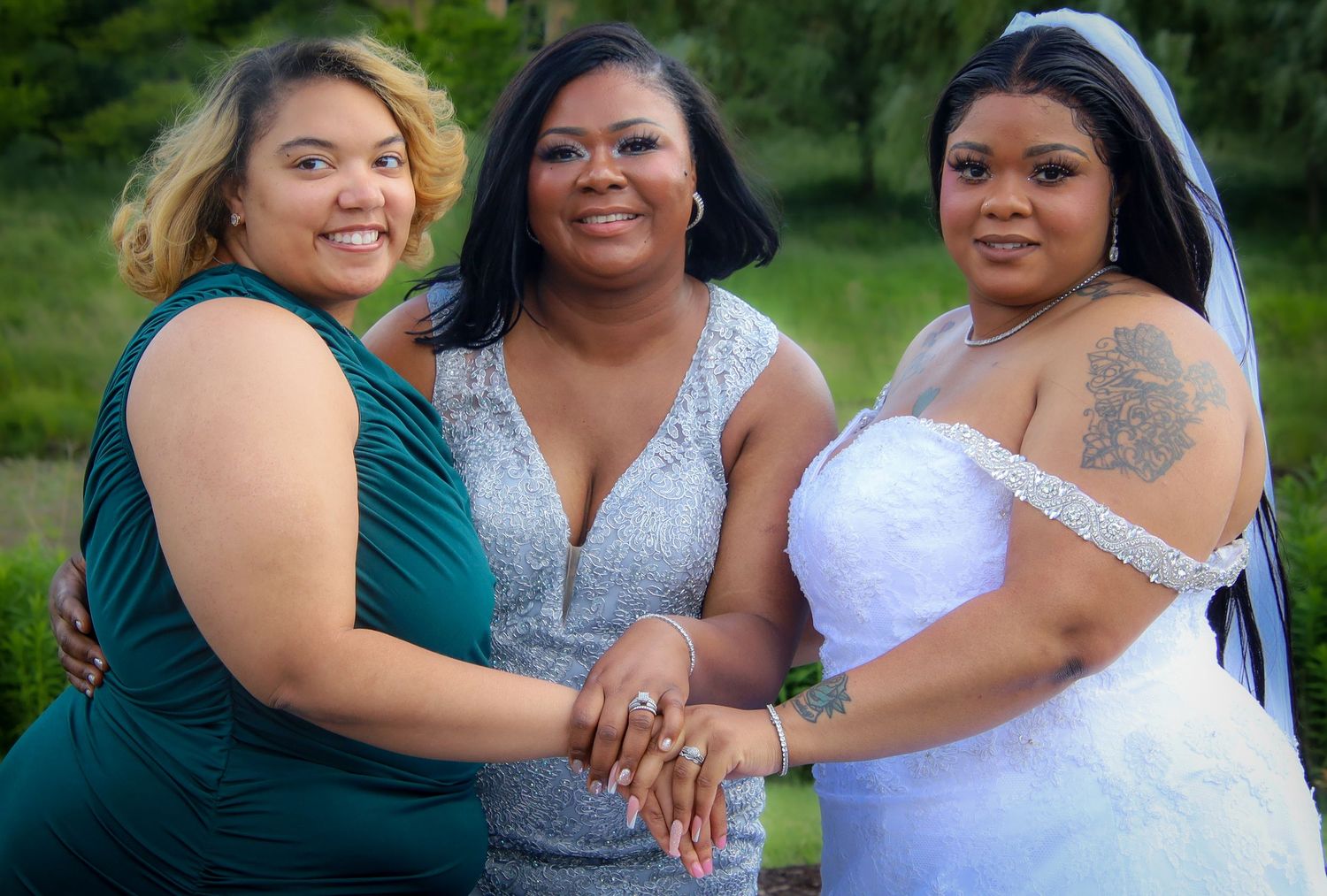 A group wearing white and silver formal dresses pose together outdoors in a garden setting.