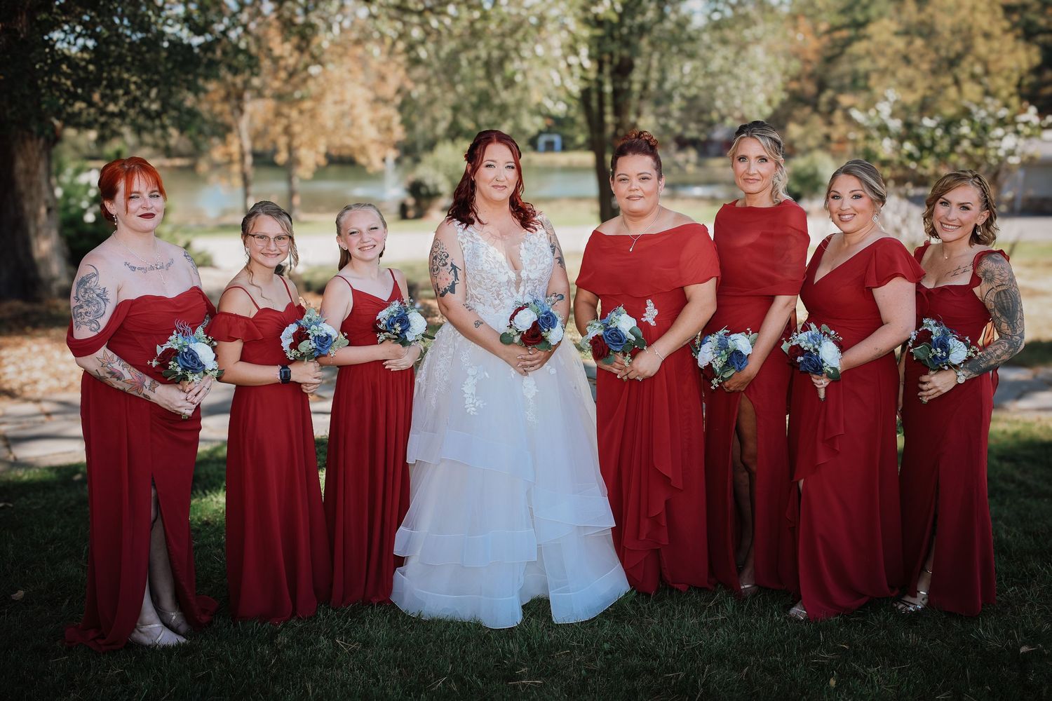 Wedding party in burgundy dresses poses outdoors with bride in white gown holding blue bouquets on autumn day.