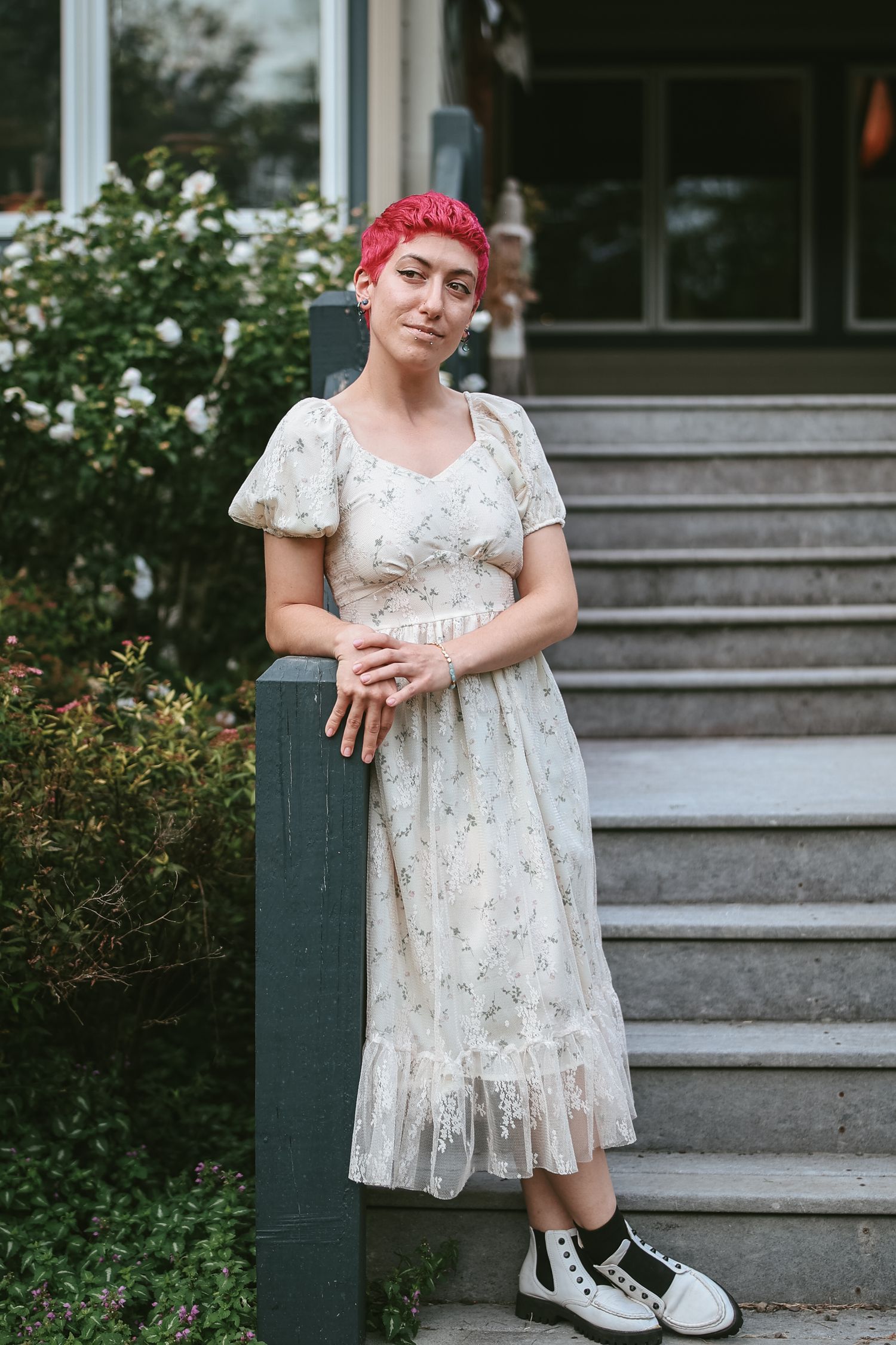 A person with bright pink hair wearing a vintage-style lace dress and sneakers stands on outdoor steps near greenery.