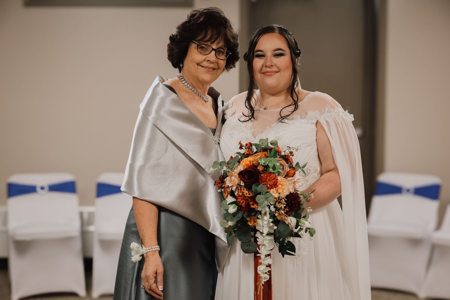 Two people pose together at a wedding ceremony, one wearing a silver shawl and one in a white dress holding a sunflower bouquet.