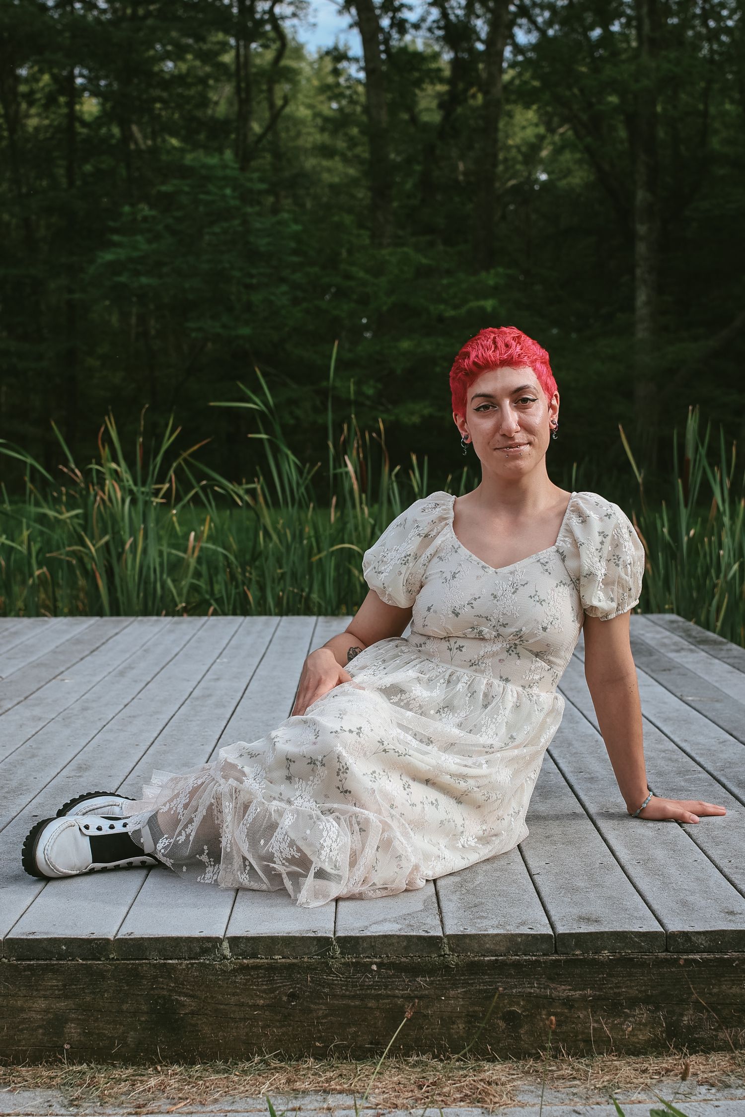 A person with bright red hair wearing a white lace dress poses playfully on a wooden deck surrounded by forest scenery.