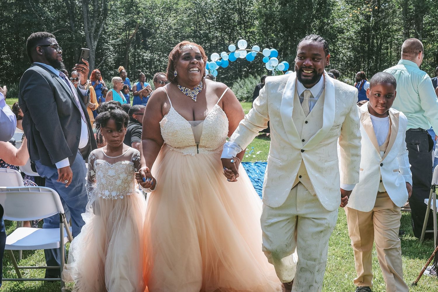 Wedding party celebrates outdoors with bridesmaids in peach dresses and groomsmen in light suits surrounded by blue balloons.