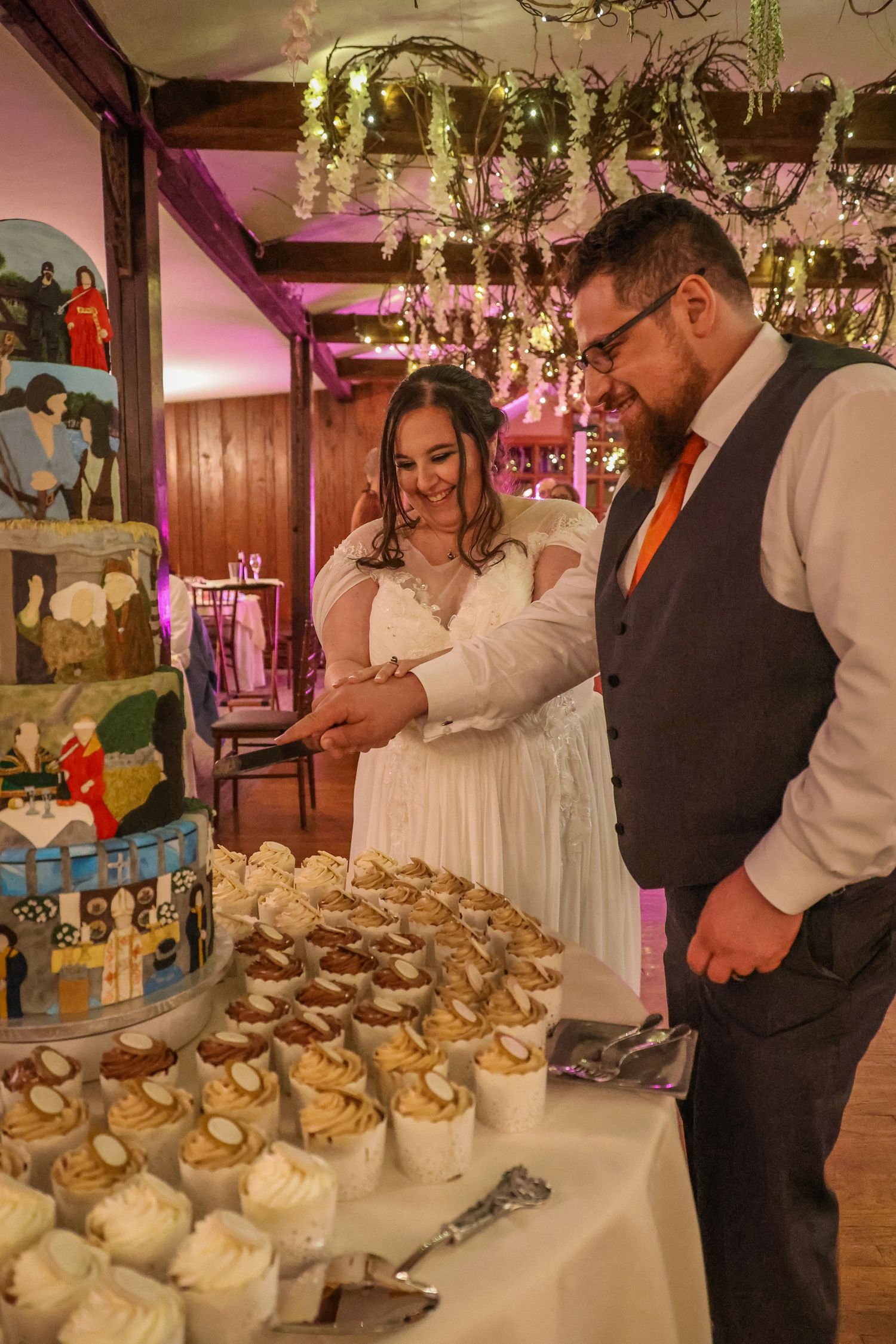 A couple cuts into their wedding cake at an elegantly decorated reception with floral arrangements and pink lighting.