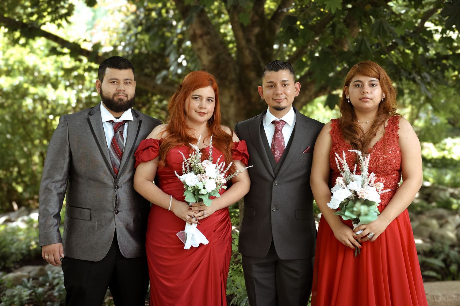 Wedding party in red dresses and gray suits poses outdoors in a garden setting with white floral bouquets.