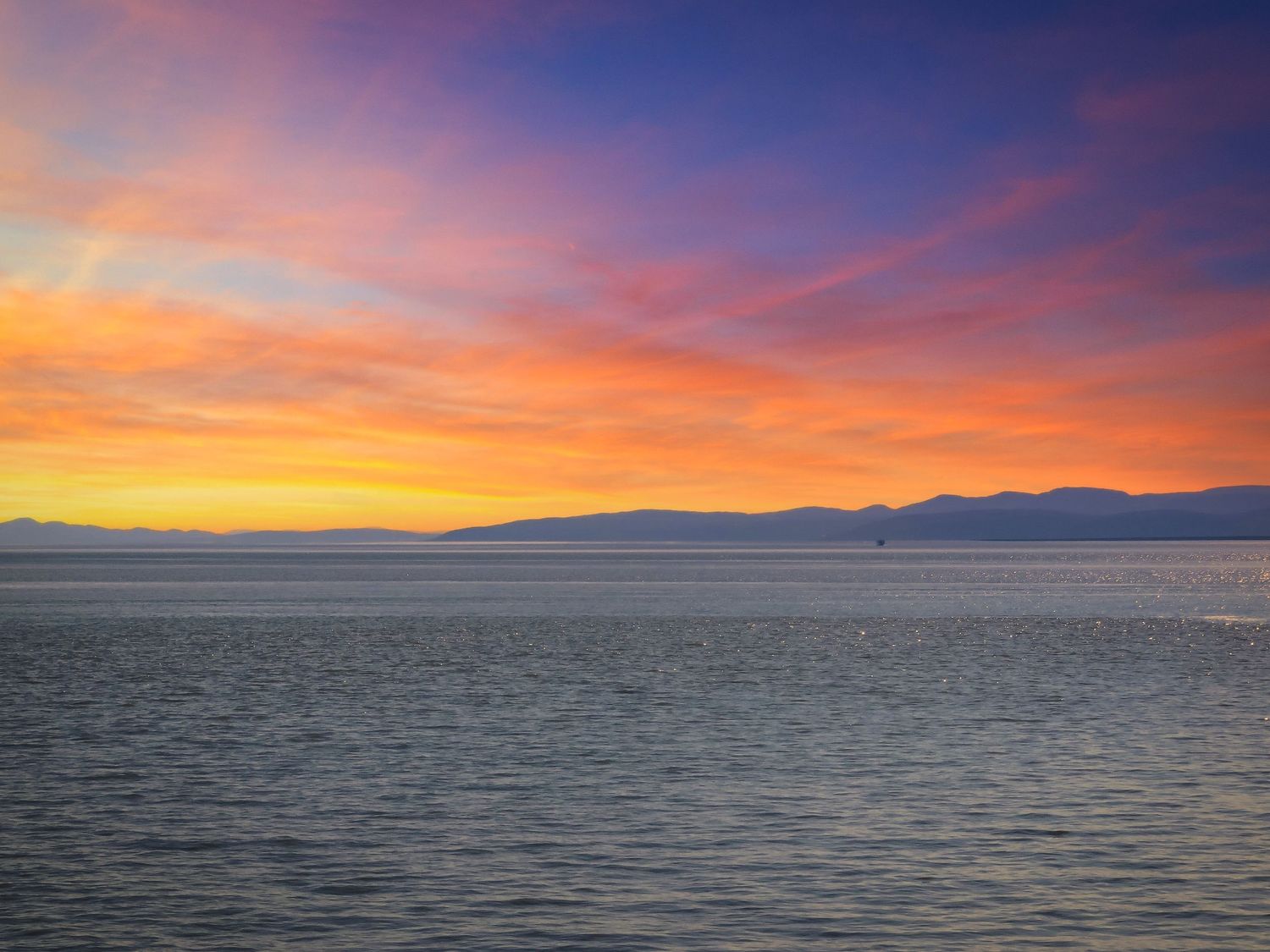 Vibrant orange and pink sunset reflects across rippling waters with mountain silhouettes in the distance.