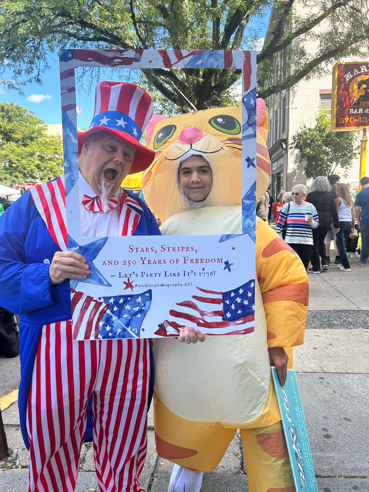 A person in patriotic red, white and blue stripes costume poses with someone in a yellow cat mascot outfit at an event.