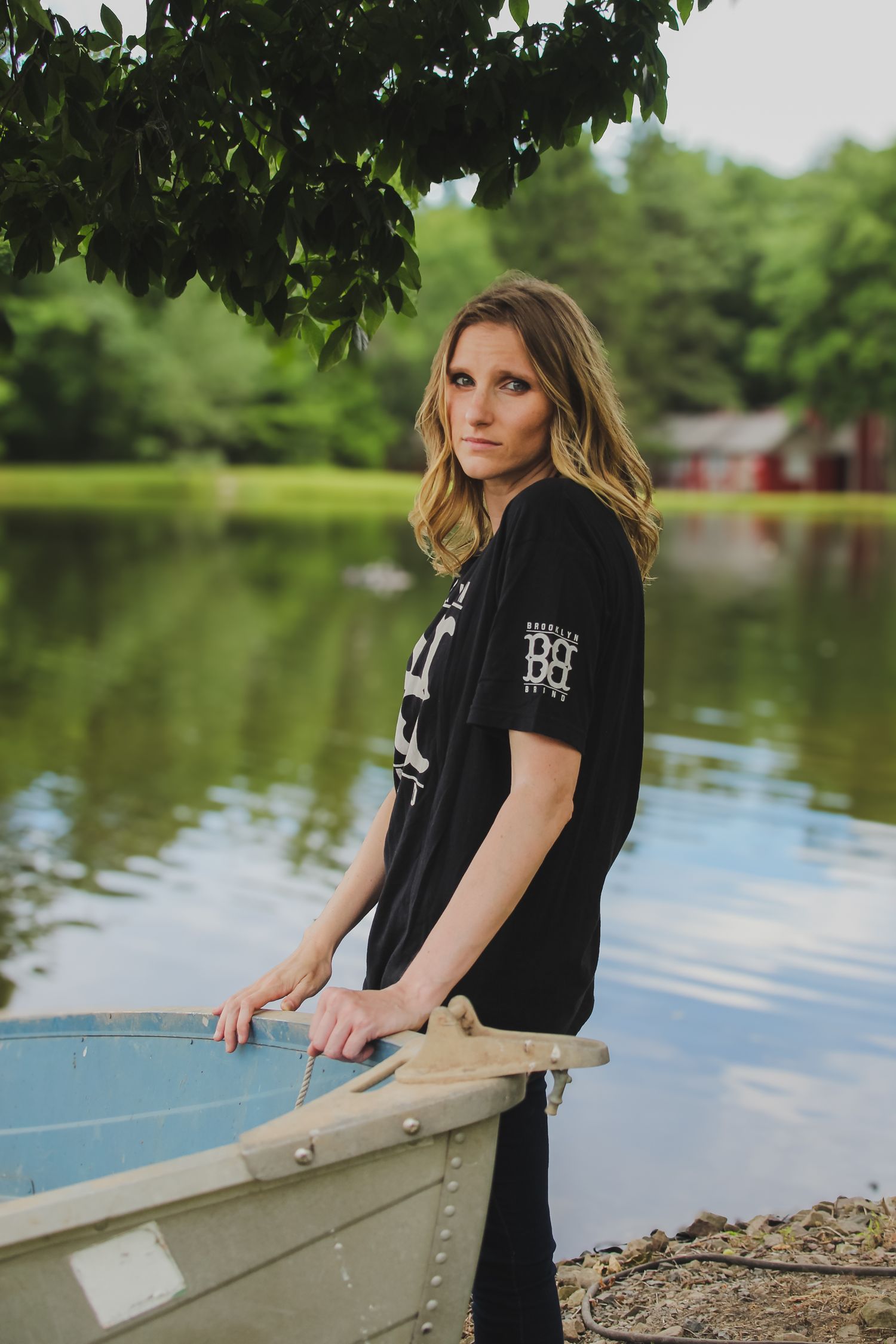 A person in a black t-shirt sits by a serene lake surrounded by greenery on a summer day.