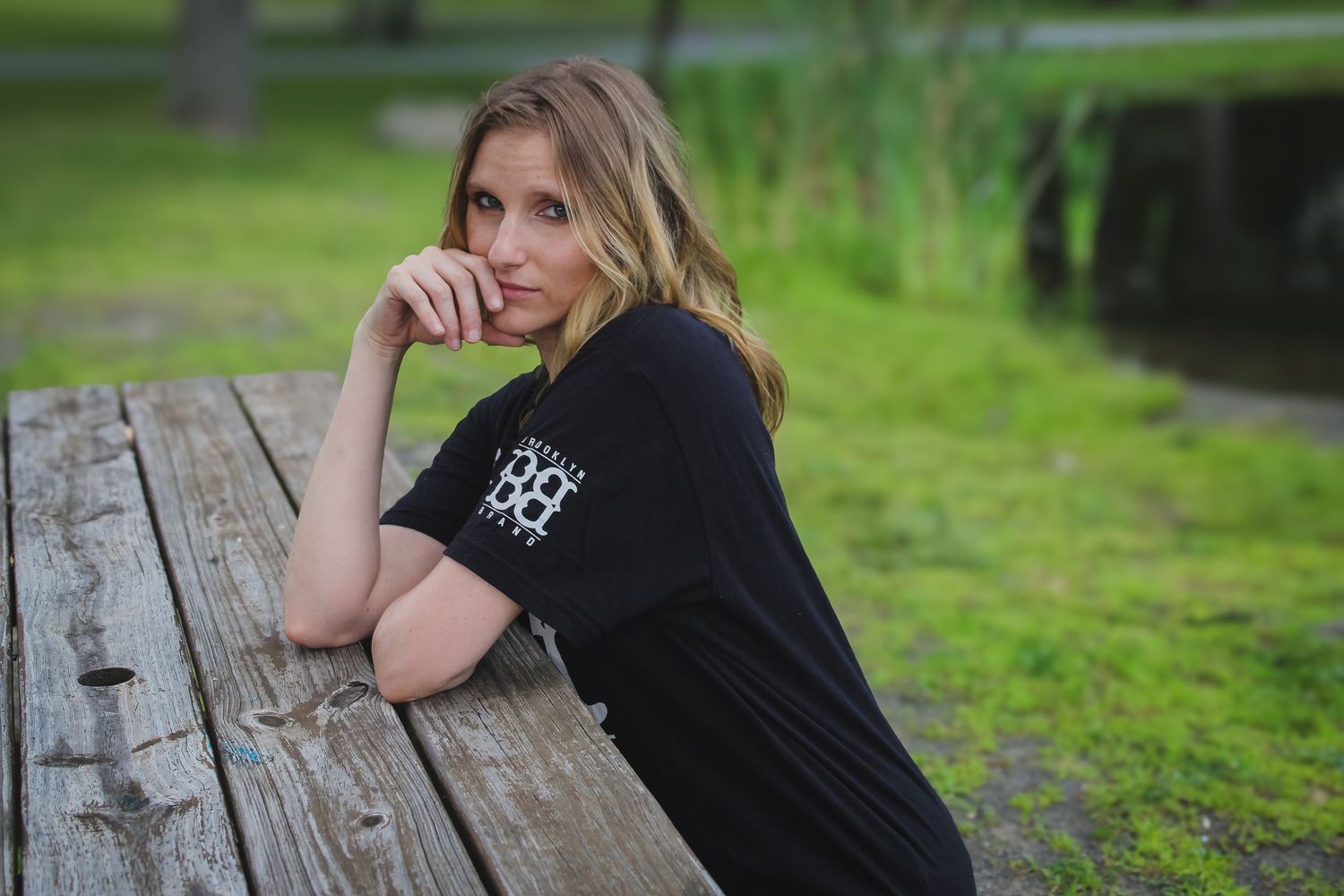 A person in black clothing sits on a wooden bench in a lush green outdoor setting.