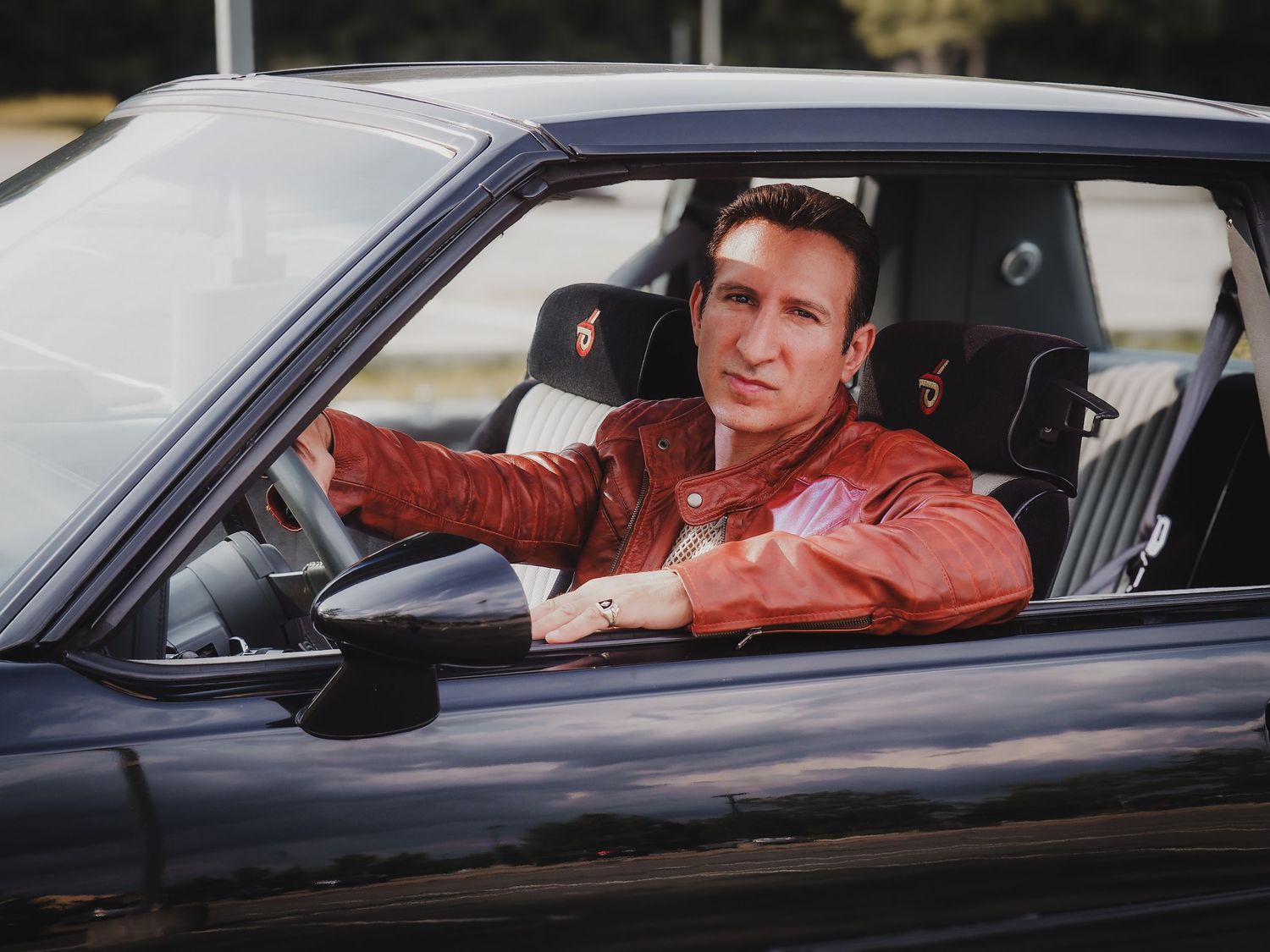 A driver wearing a brown leather jacket sits at the wheel of a dark-colored car.
