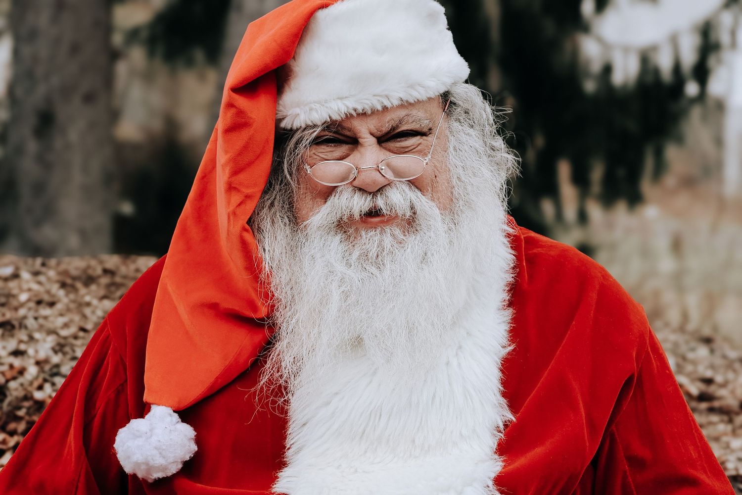 Santa Claus in traditional red suit and white beard smiling while sitting outdoors in a winter setting.