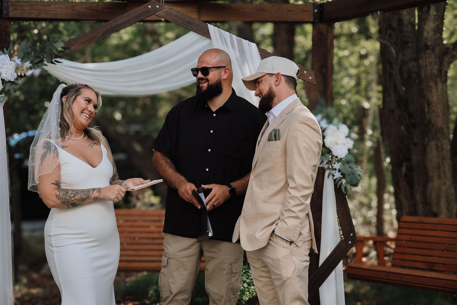 An outdoor wedding ceremony takes place under a white draped arch in a wooded setting.