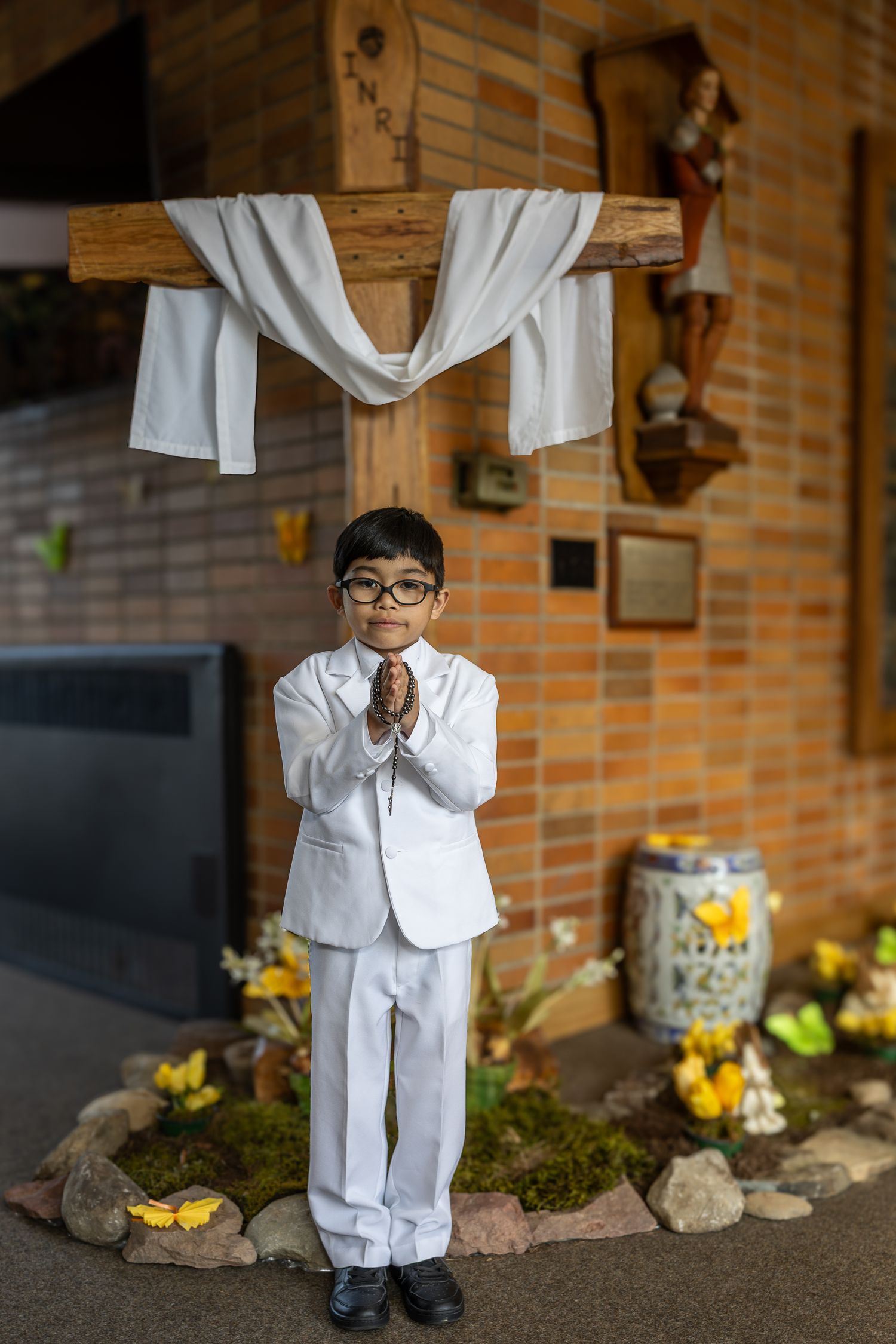 A small child wearing a white lab coat and glasses stands against a brick wall with decorative garden elements.
