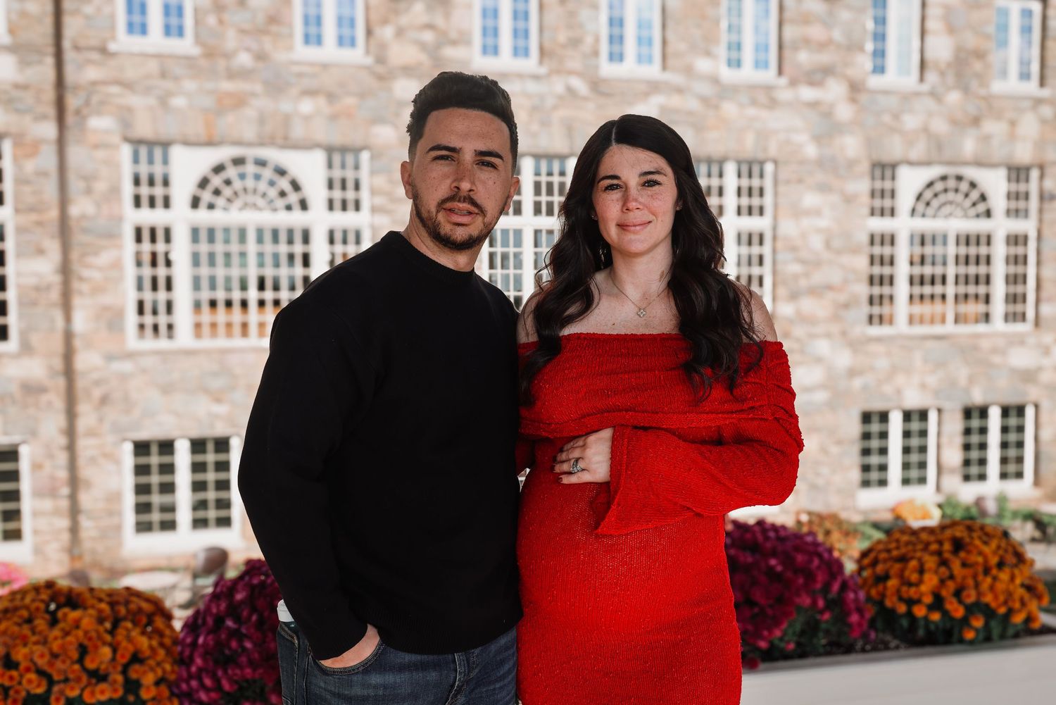 A couple in coordinating black and red outfits pose together in front of a stone building with fall flowers.