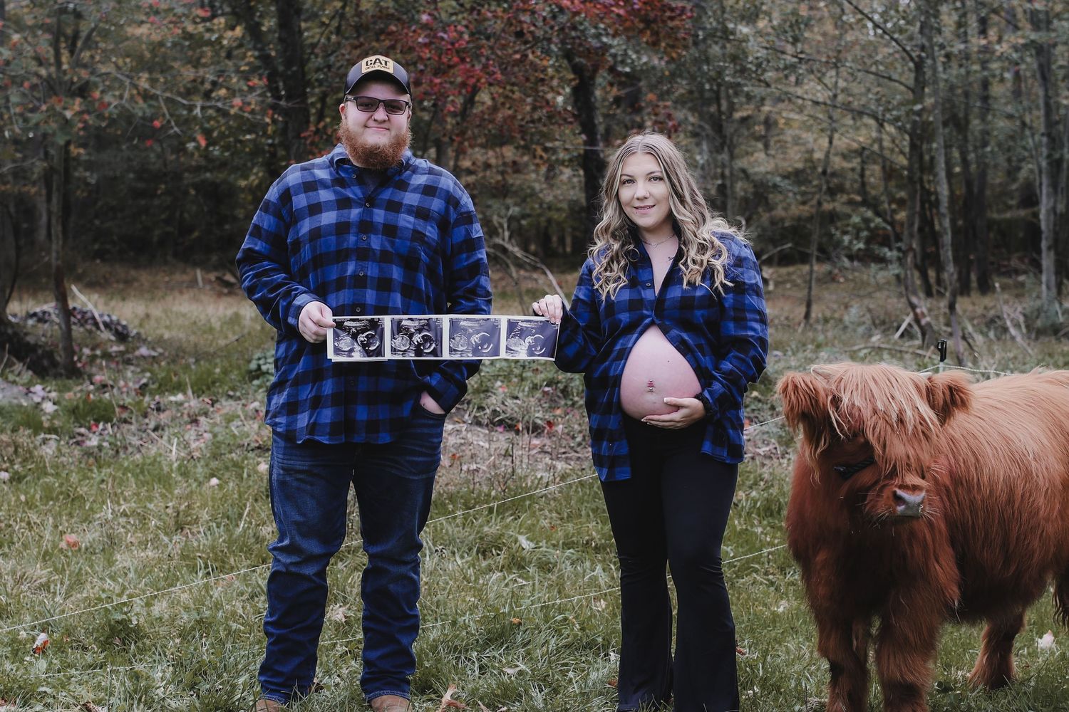 A couple stands in a wooded area with their brown horse while looking at their phones.