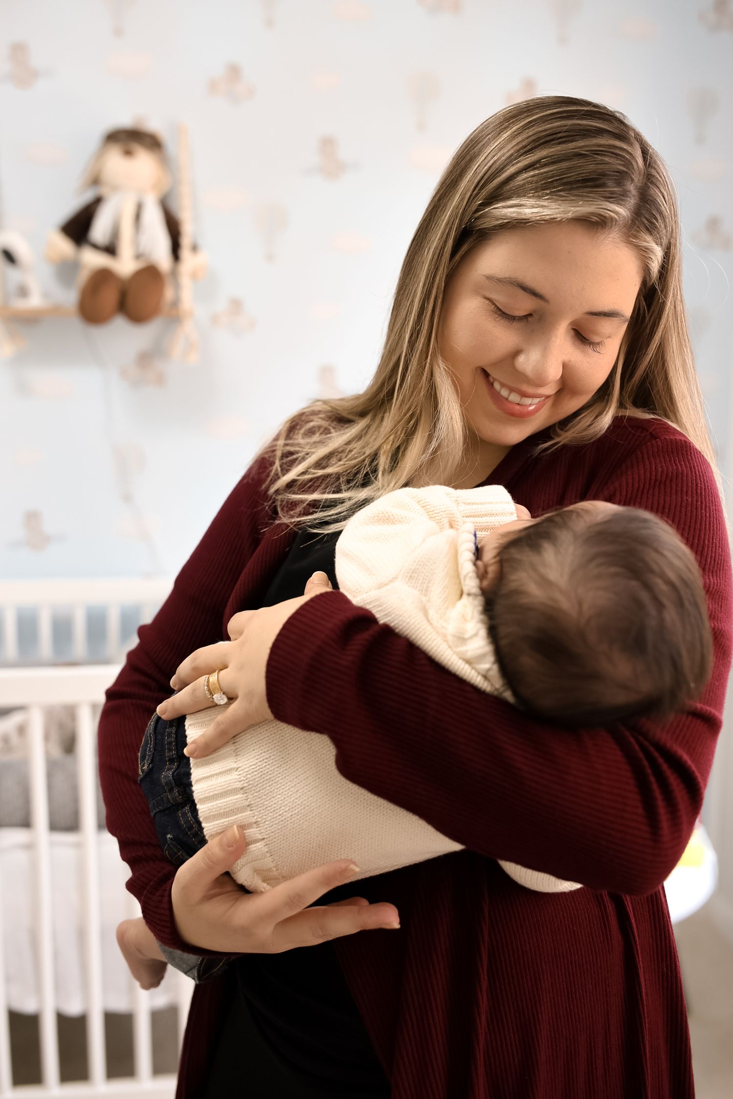 A parent wearing a burgundy sweater holds and smiles at their baby in a white onesie in a nursery room.