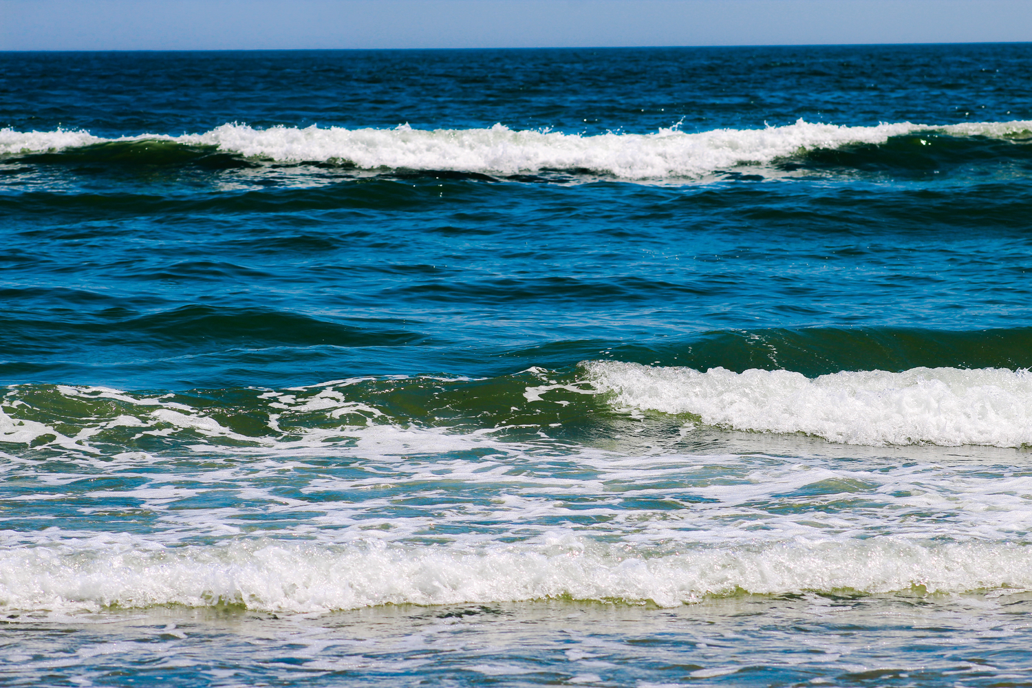 Ocean waves with white foam crashing on a sunny beach, featuring deep blue water and clear skies.
