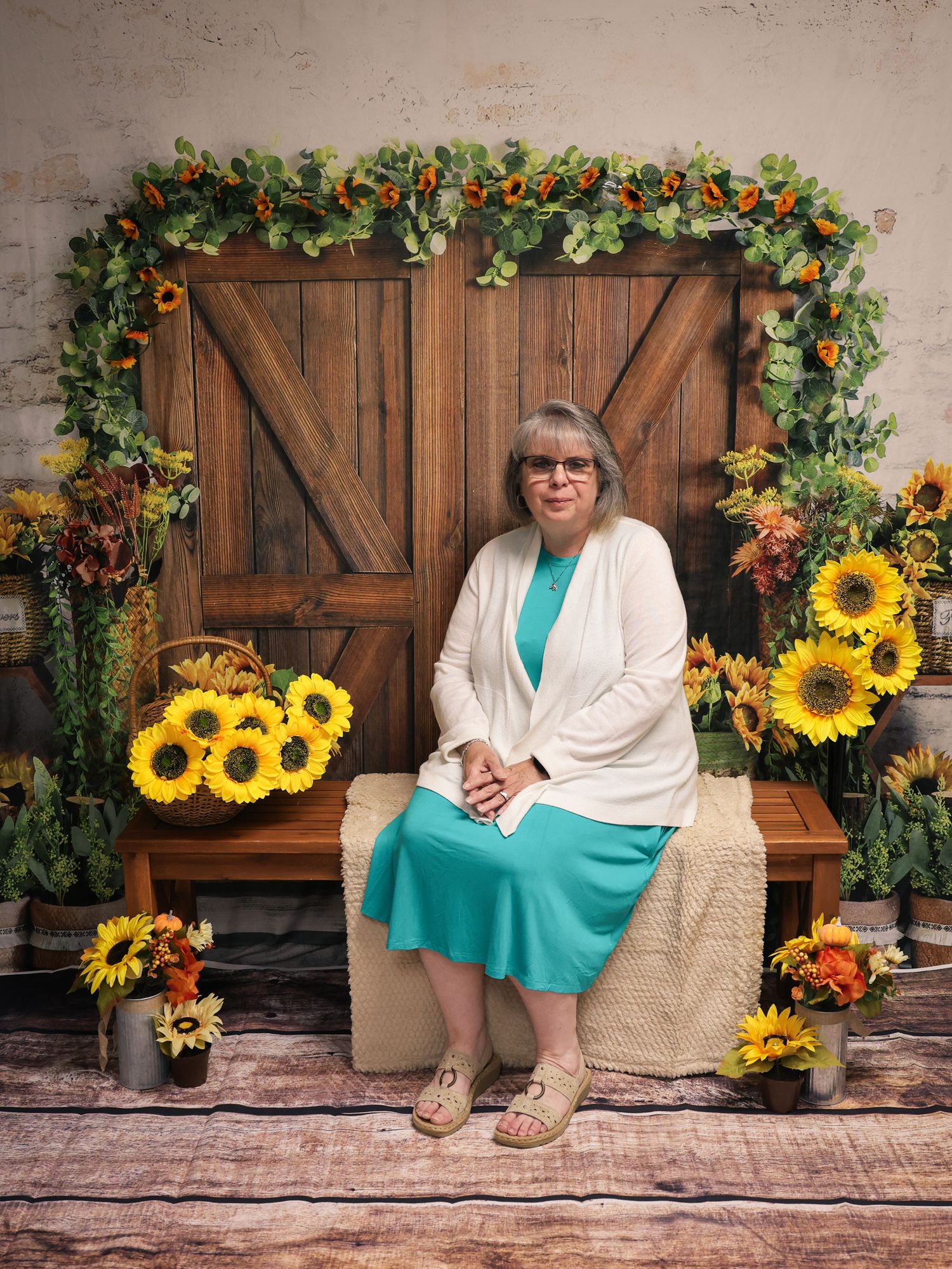 Woman in teal dress and white cardigan sits on bench surrounded by sunflowers and rustic barn door backdrop.