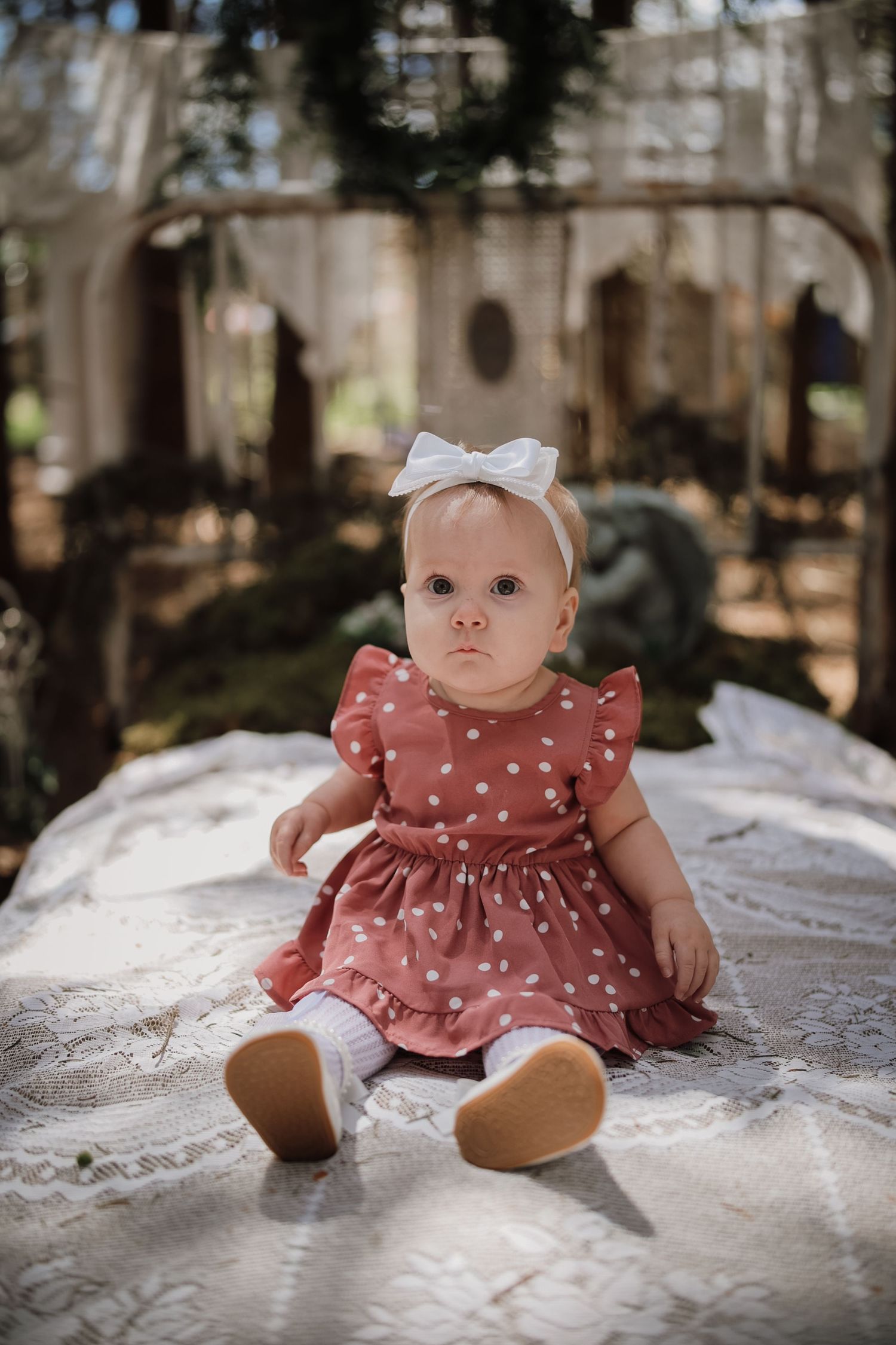 Baby girl in pink polka dot dress and white bow headband sitting on lace blanket outdoors.