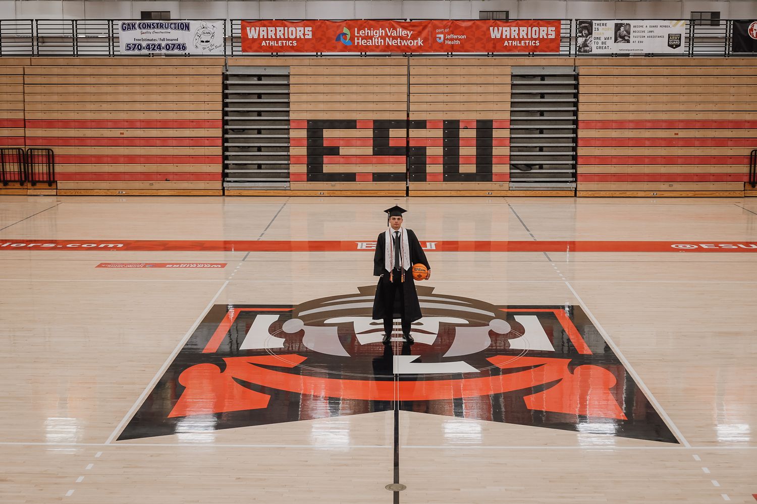 Graduate in cap and gown stands at center court in ESU Warriors gymnasium with bold red and black logo.