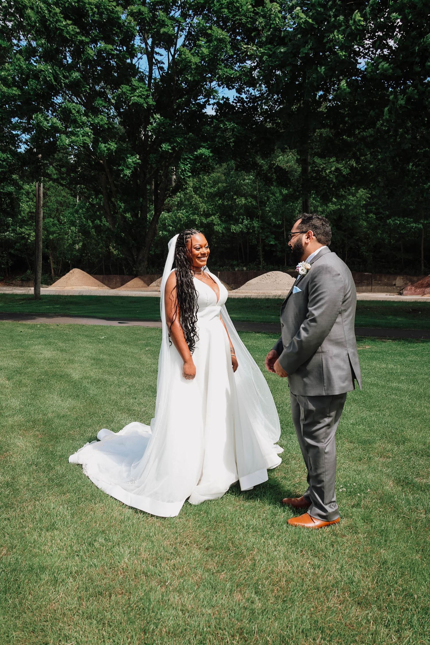 Bride in a flowing white gown with veil smiling at groom in gray suit on a lush green lawn.