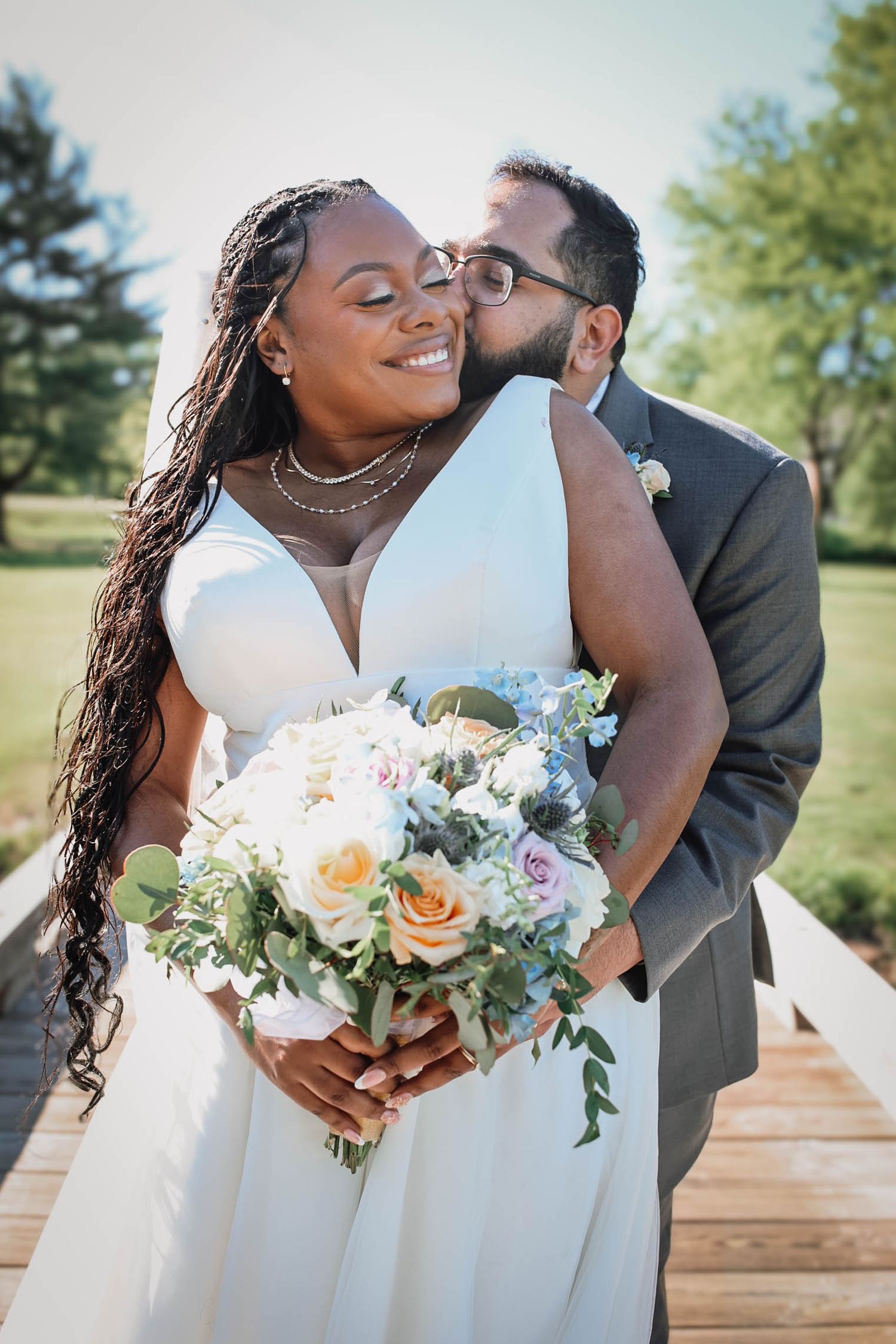 A bride in a white gown holds a pastel bouquet as her groom kisses her cheek on a sunny outdoor wedding day.