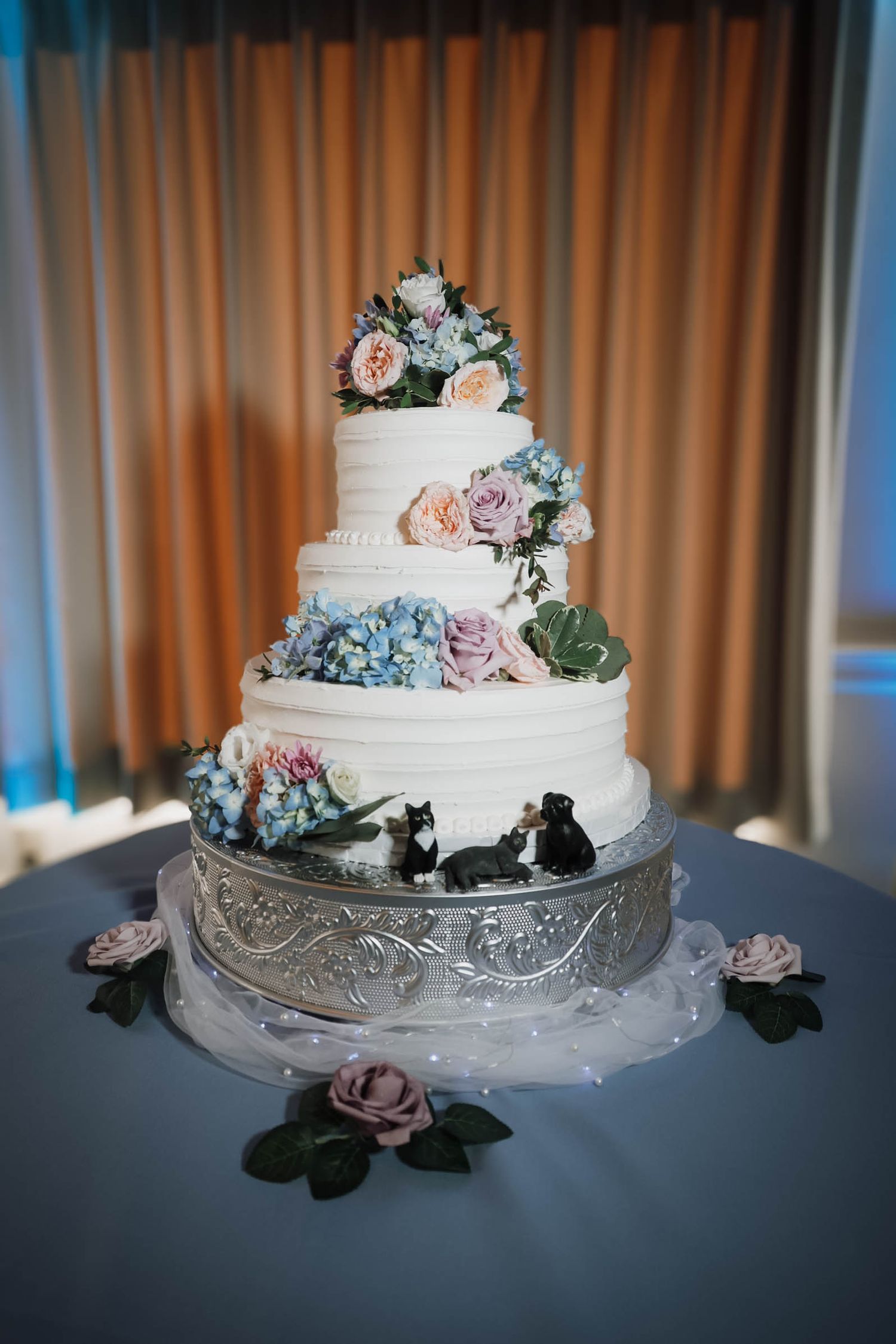 Four-tier white wedding cake with pastel flowers, cat toppers, on silver stand.
