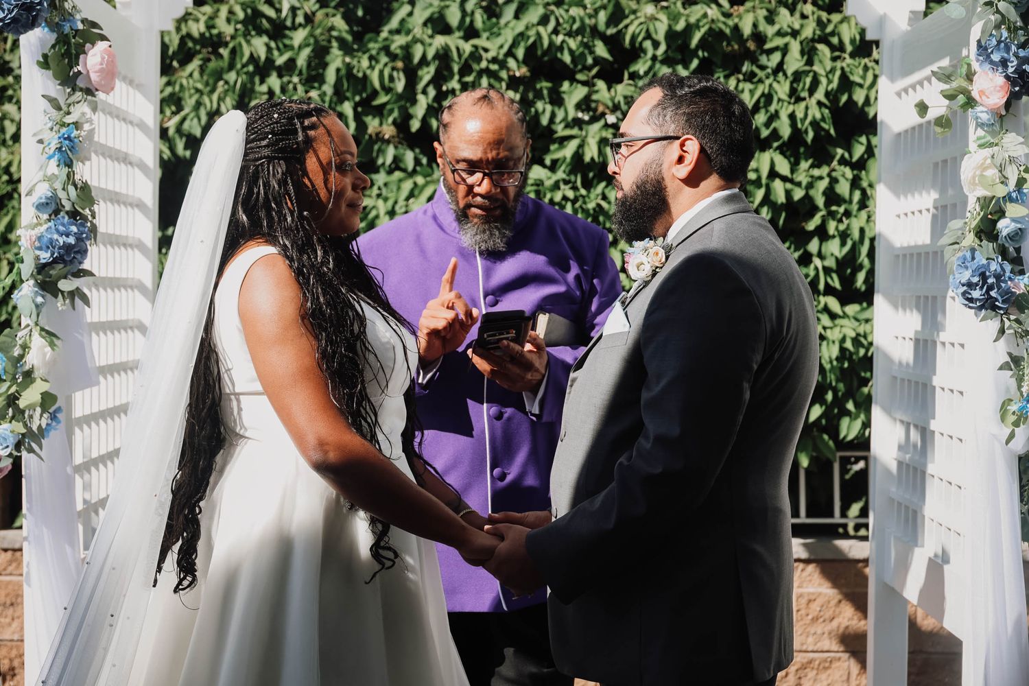 Bride and groom exchanging vows outdoors with officiant in purple robe at floral arch ceremony.
