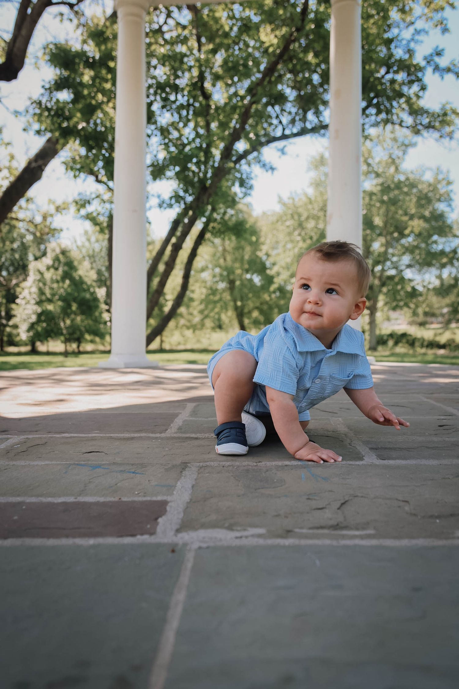 A baby boy in a blue outfit crawls on a stone patio surrounded by white columns and green trees.