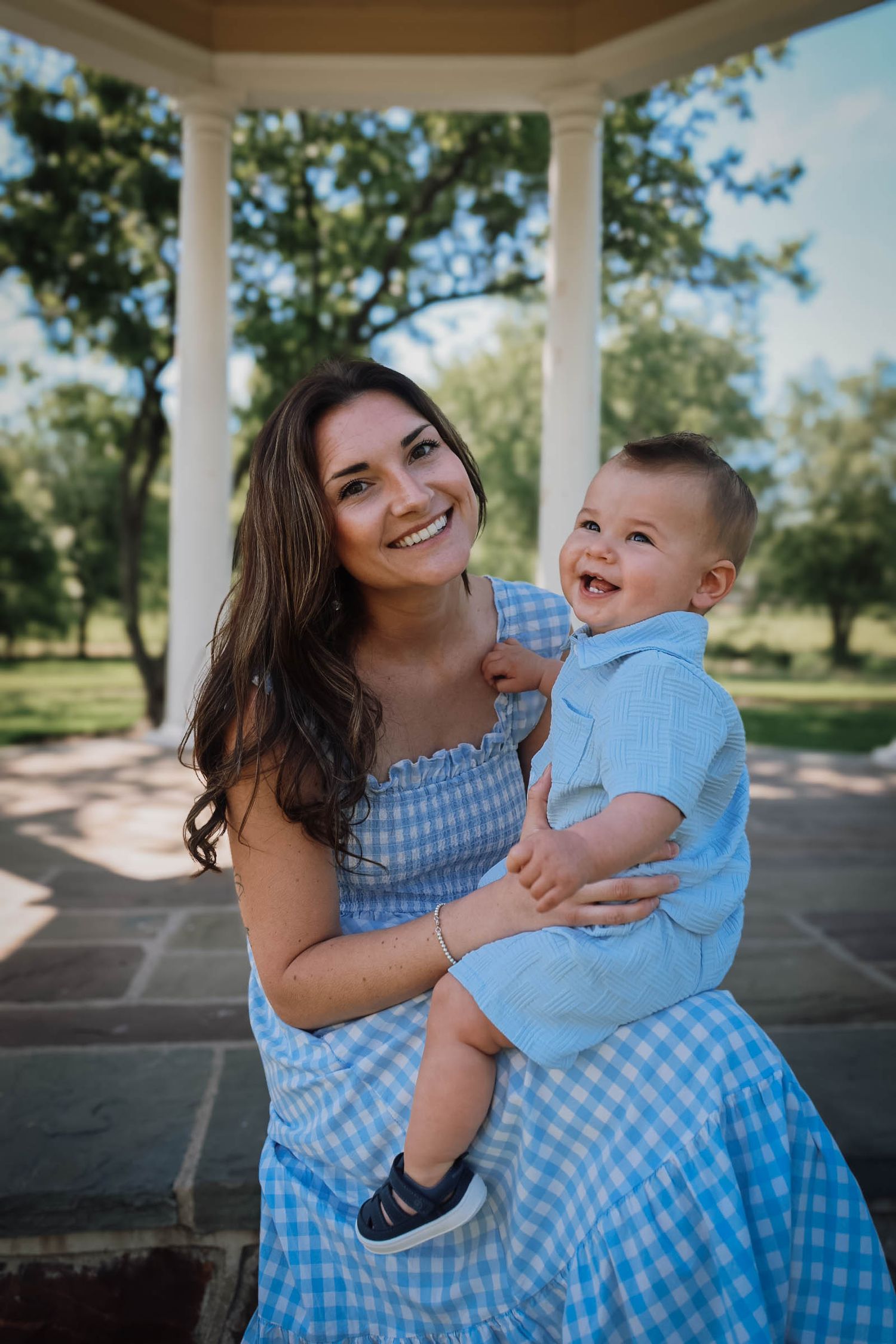 Smiling mother in blue gingham dress holds her laughing baby boy under a park gazebo.