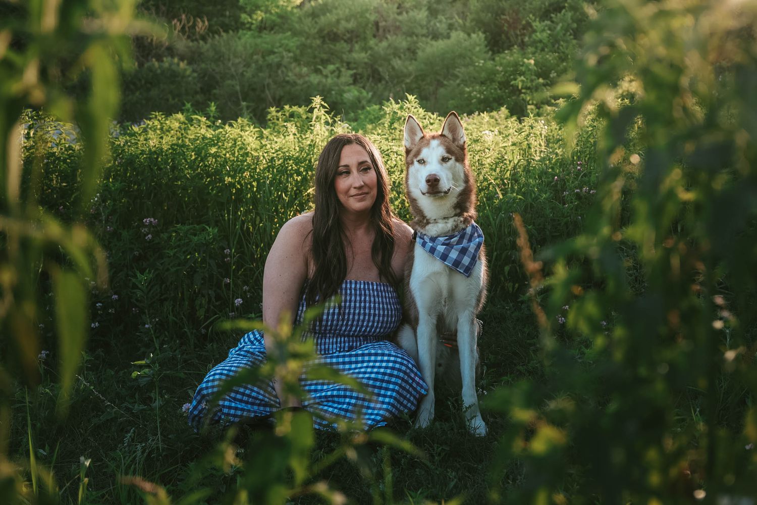 Woman in blue gingham dress sits with her Husky dog wearing a bandana in a lush green meadow.