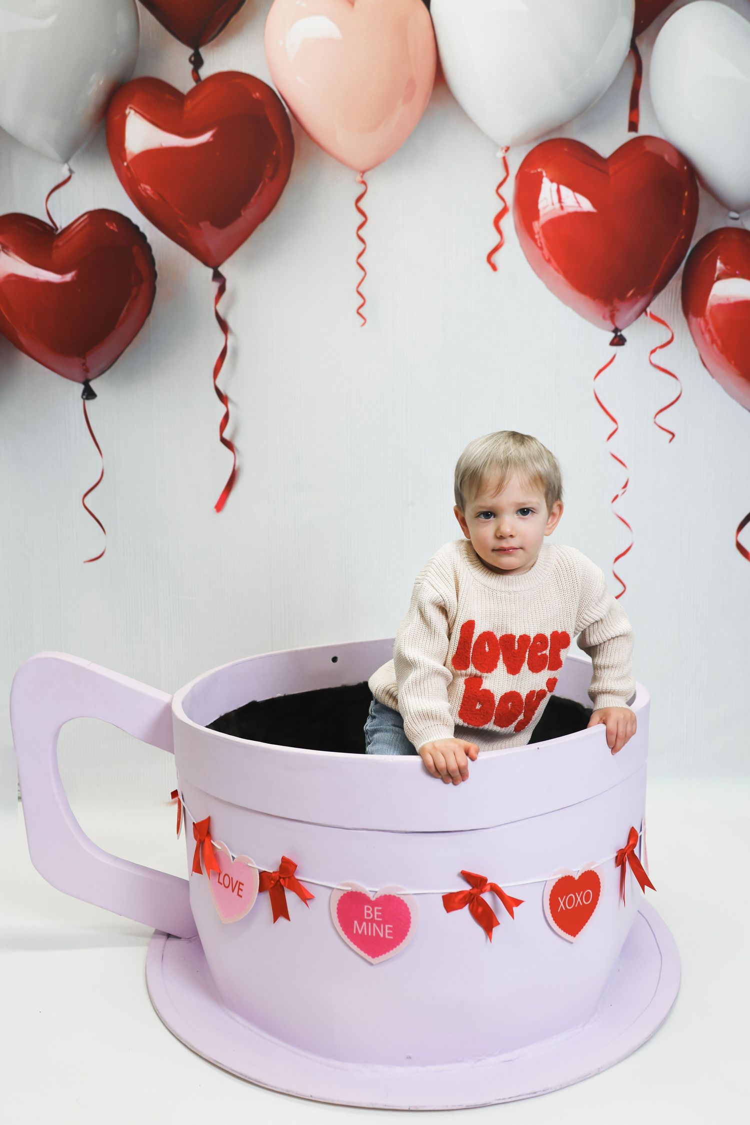 Toddler sitting in giant teacup prop surrounded by red and pink heart balloons for Valentine's Day photo.