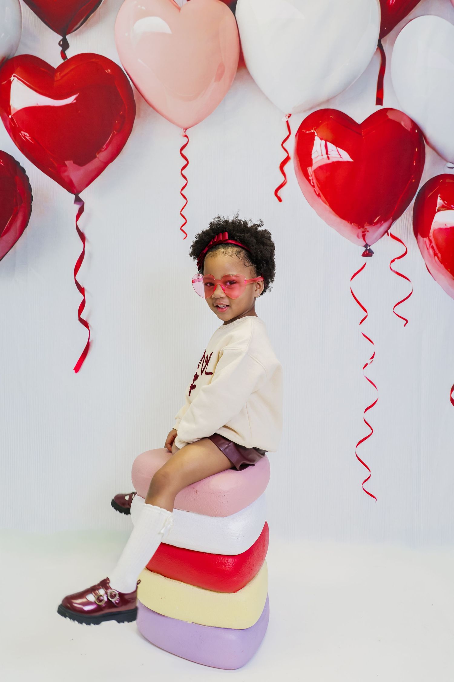 Young girl in pink sunglasses sits on colorful stacked cushions surrounded by red, pink, and white heart-shaped balloons.
