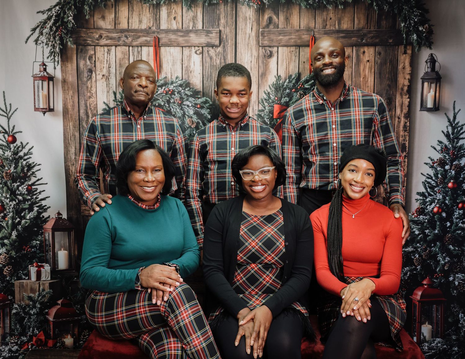 A joyful Black family of six poses for a festive Christmas portrait in matching plaid outfits before a rustic holiday backdrop.