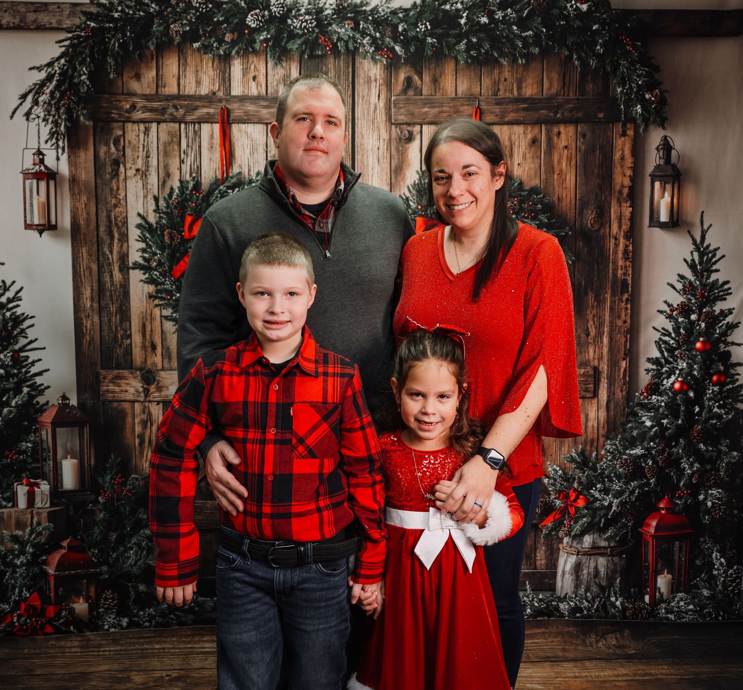 Family of four poses for a festive Christmas portrait with holiday decorations and Christmas trees in background.