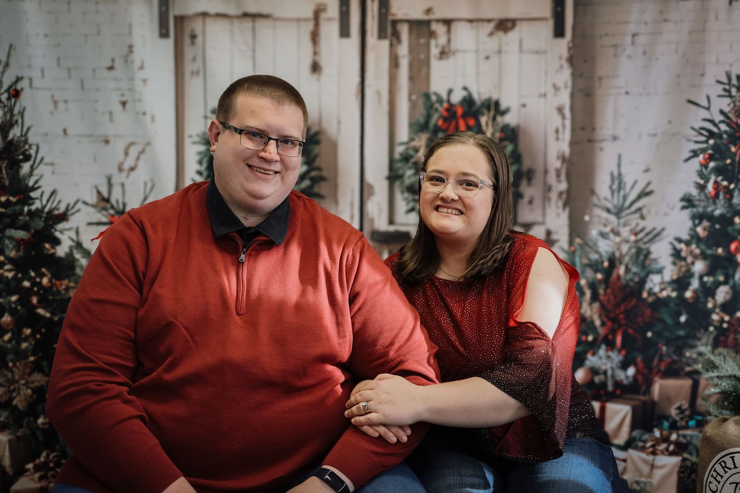 Couple in matching red sweaters posing together in front of Christmas trees and holiday decorations.