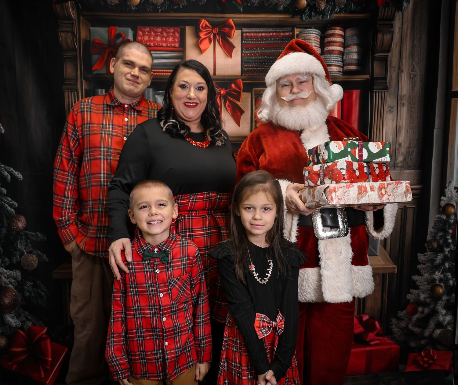 Family of four poses with Santa Claus holding gifts in a festive Christmas-themed photo studio setting.