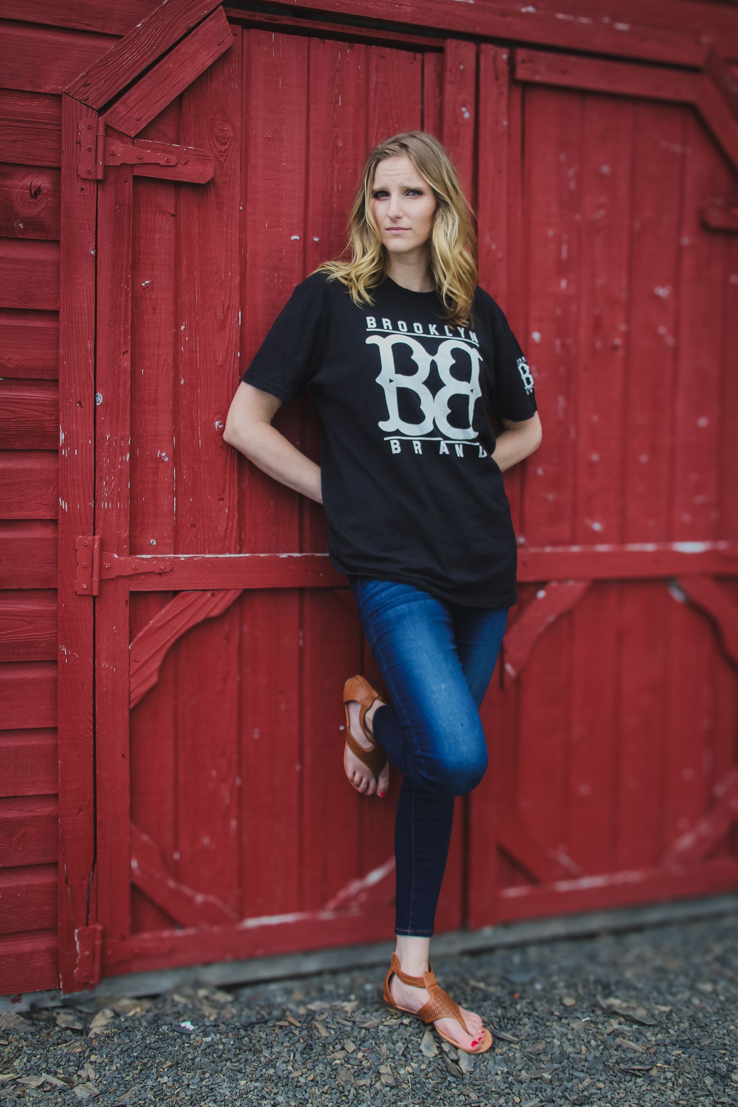 Fashion model in black graphic t-shirt and jeans poses against vibrant red barn wall in outdoor photoshoot.