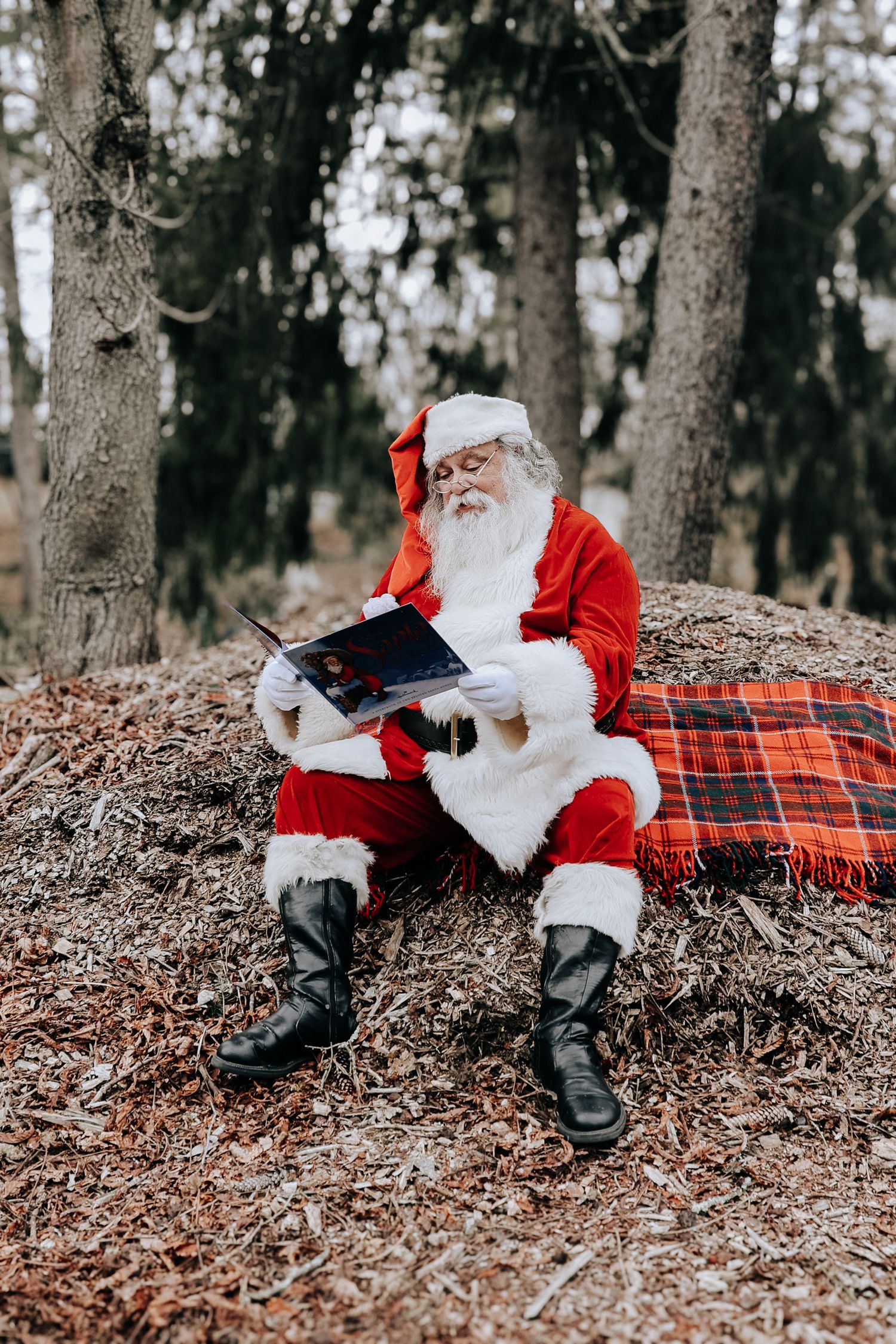 Santa Claus relaxes on a plaid blanket in the woods while reading letters on a winter day.