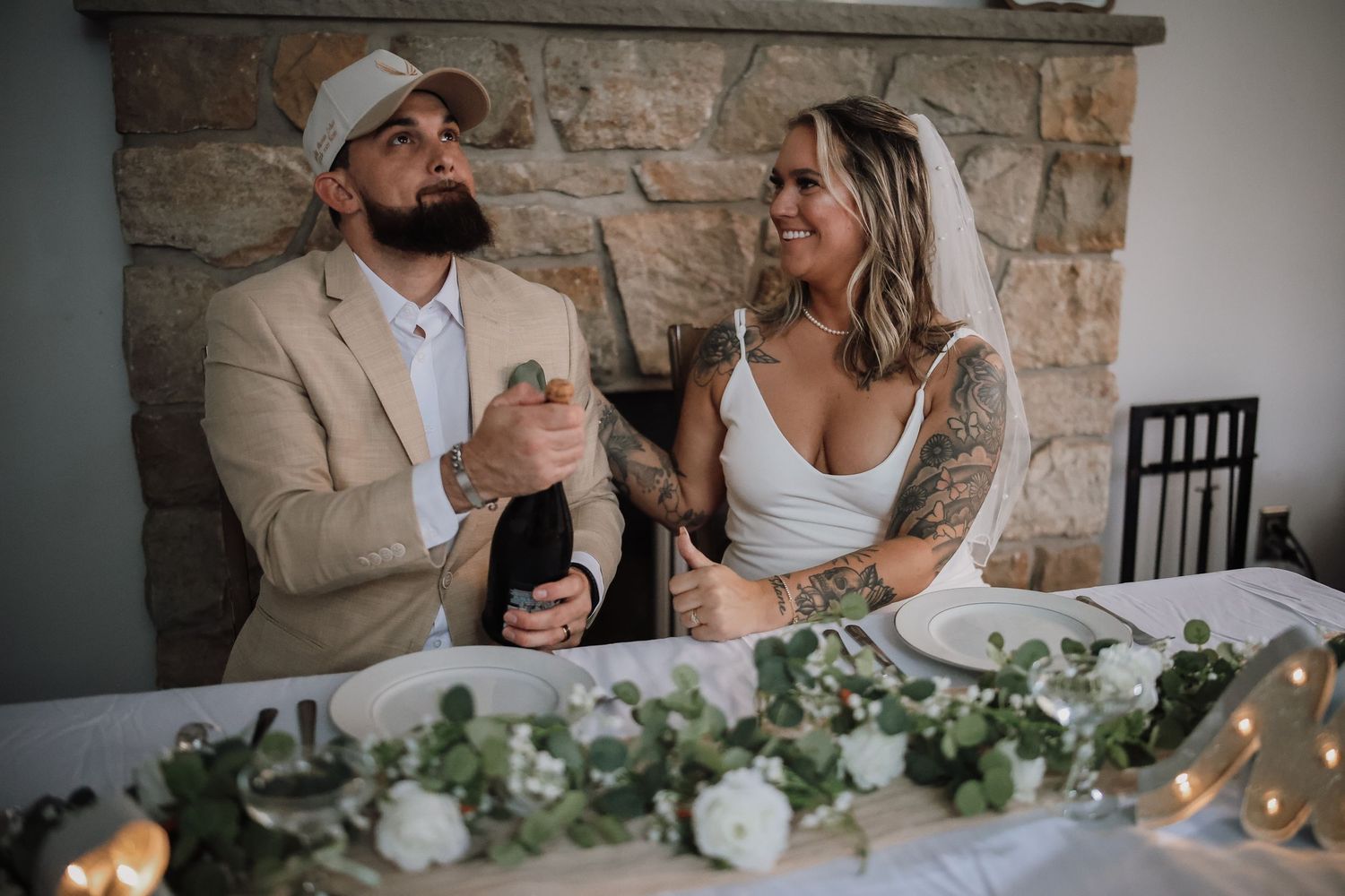 Couple sits at elegant rustic wedding reception table decorated with greenery and white floral centerpieces.