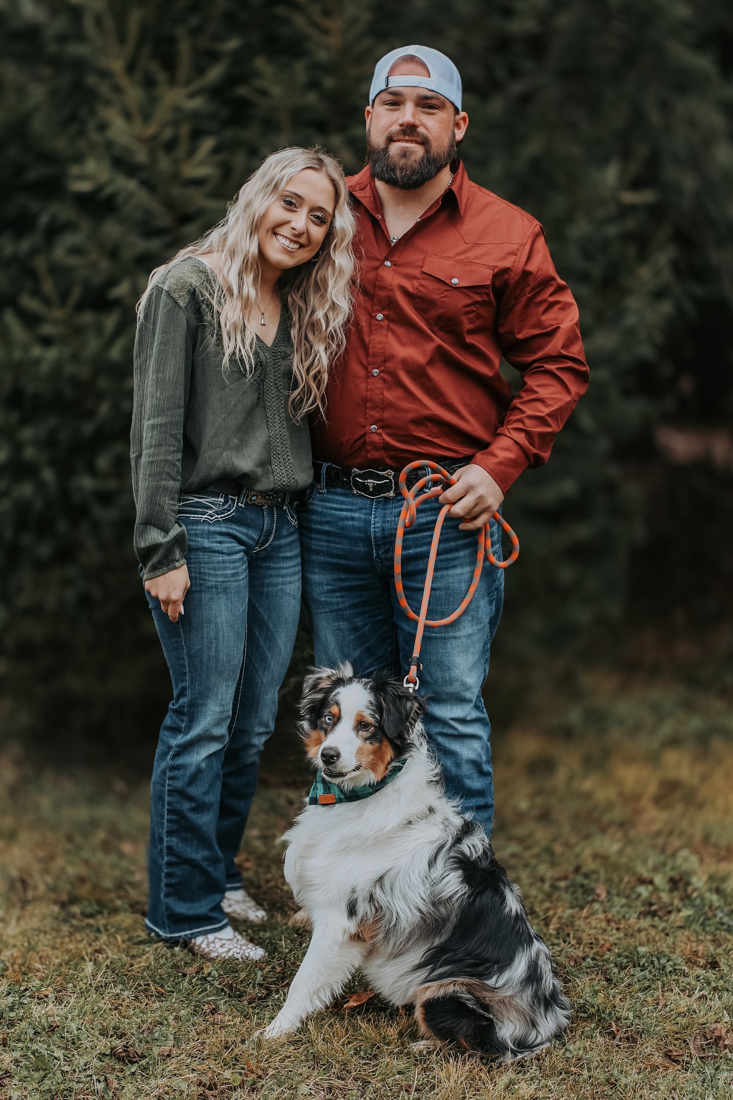 Couple in casual outdoor wear poses with their Australian Shepherd dog against a forest backdrop.