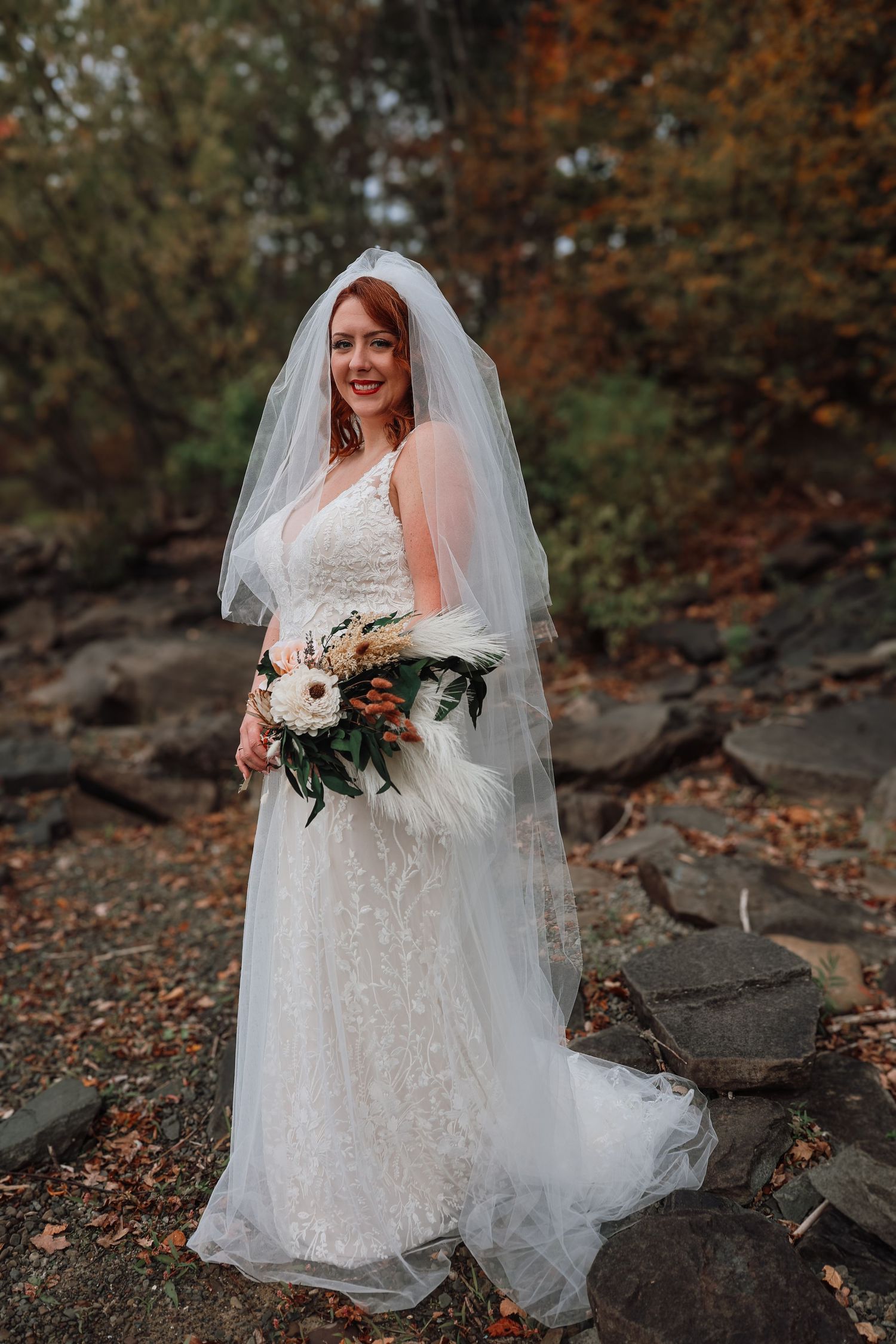 A bride in a lace wedding dress poses outdoors among autumn leaves and rocks holding a white and burgundy bouquet.