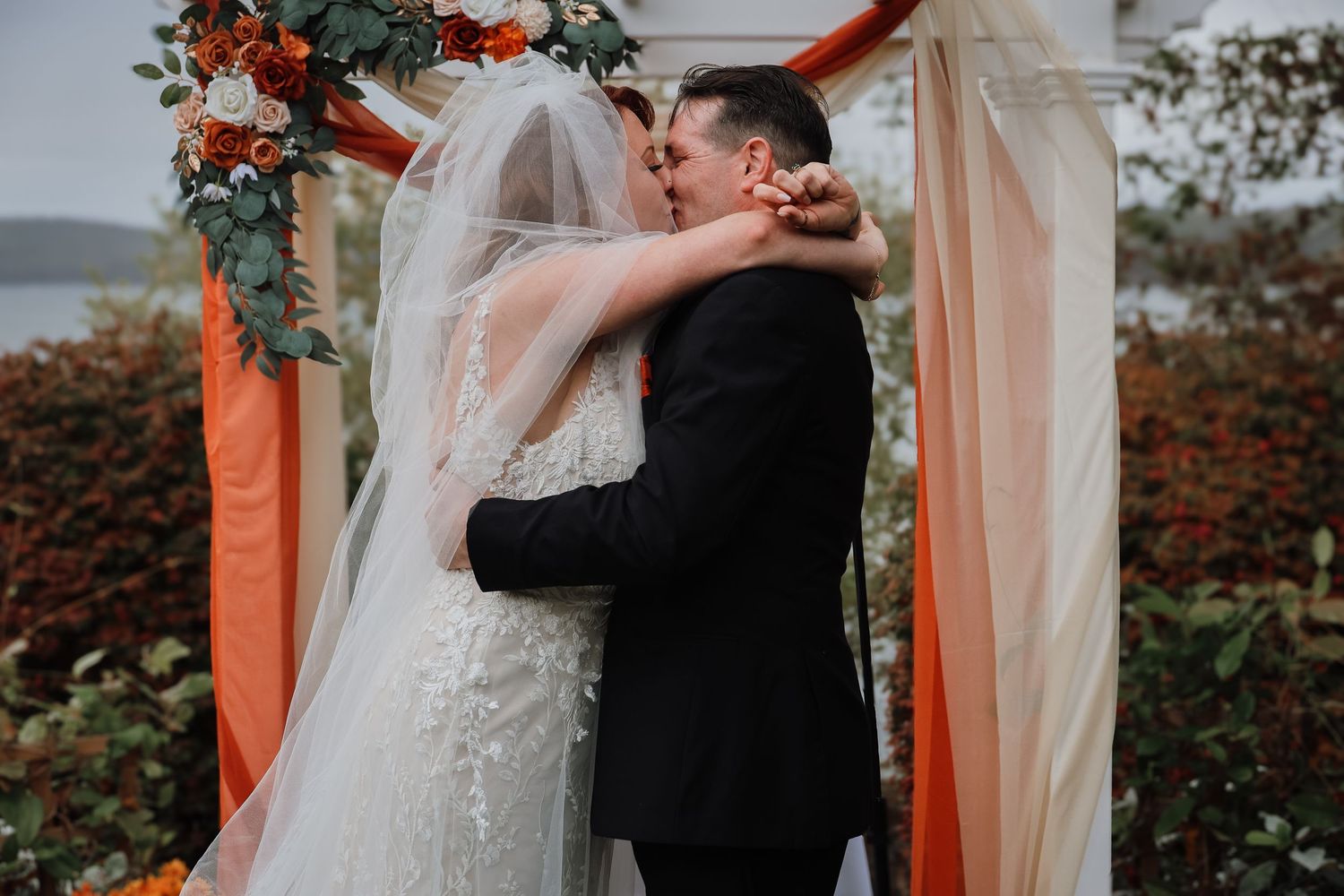 A romantic wedding ceremony moment under a floral arch with coral and peach draping by the water.