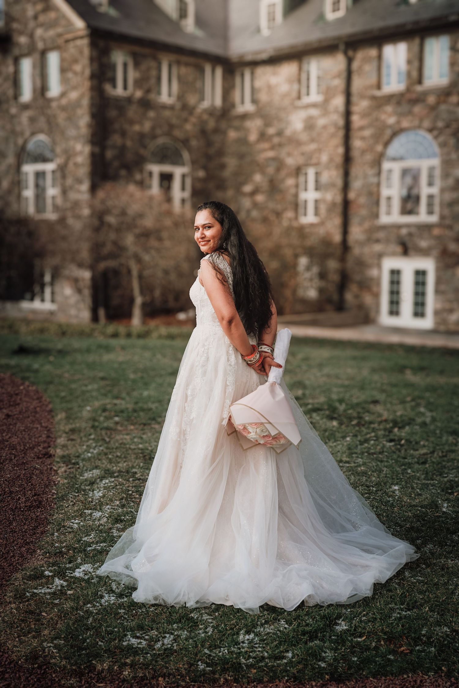 A bride in a flowing white gown twirls gracefully on a lawn in front of a stone building.