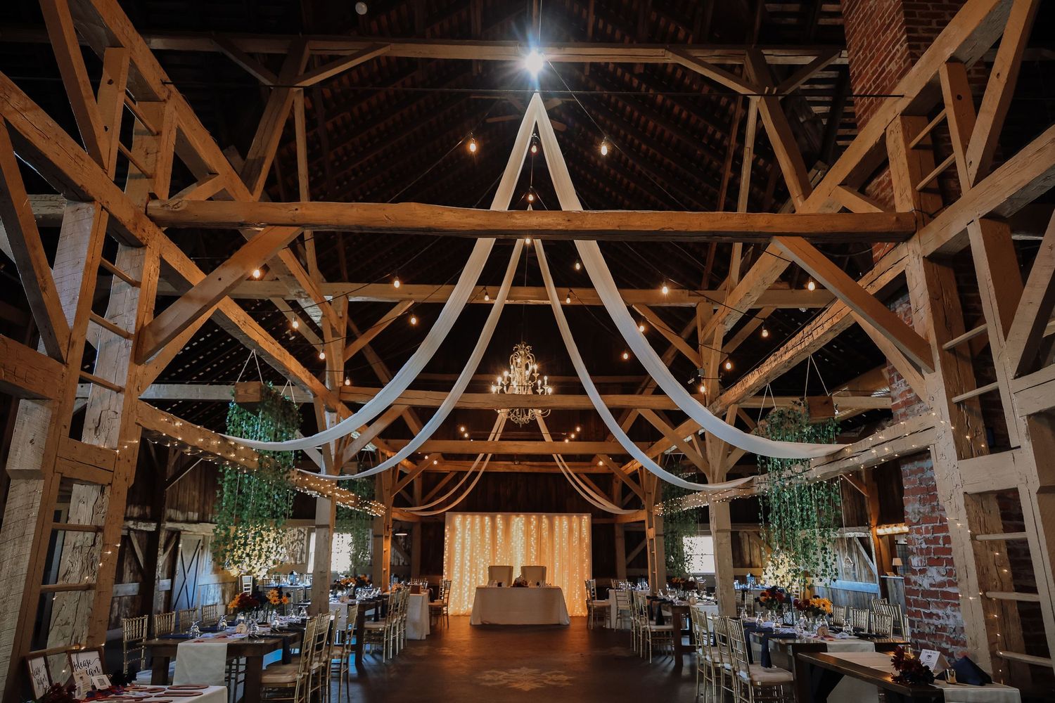 Rustic barn wedding venue interior decorated with string lights and chandeliers hanging from wooden beams.