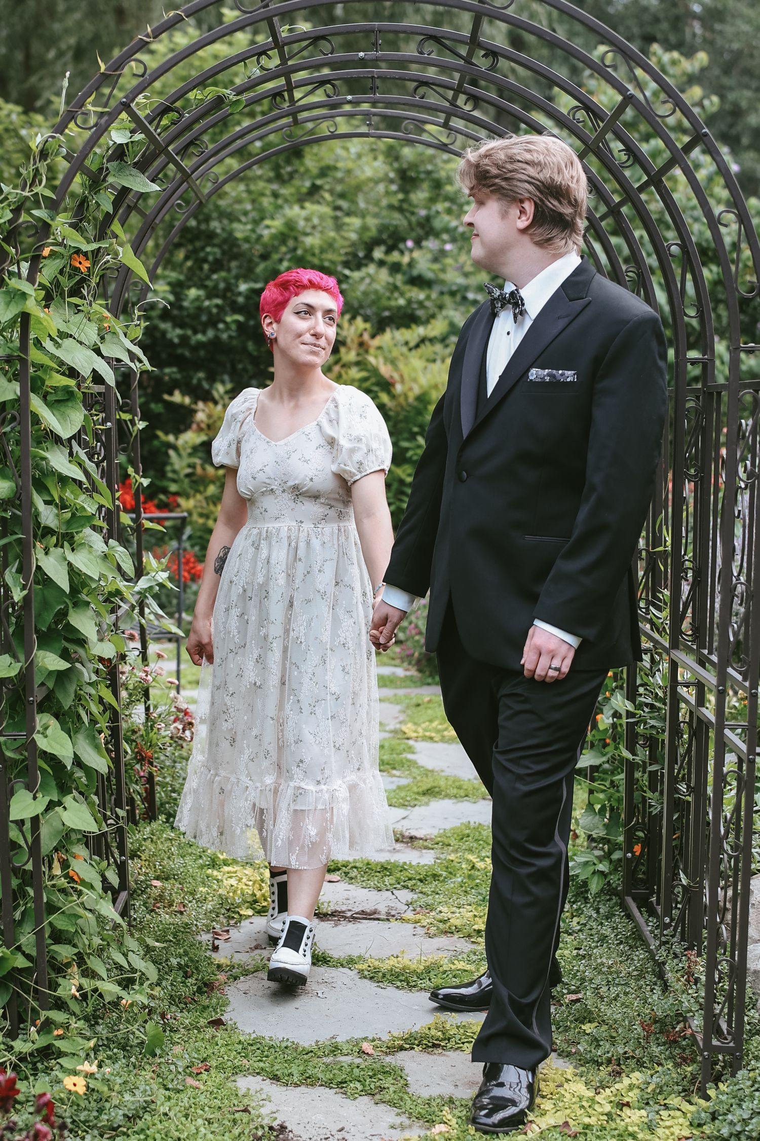 A couple walks together through a garden archway with lush greenery and flowers during a wedding photoshoot.